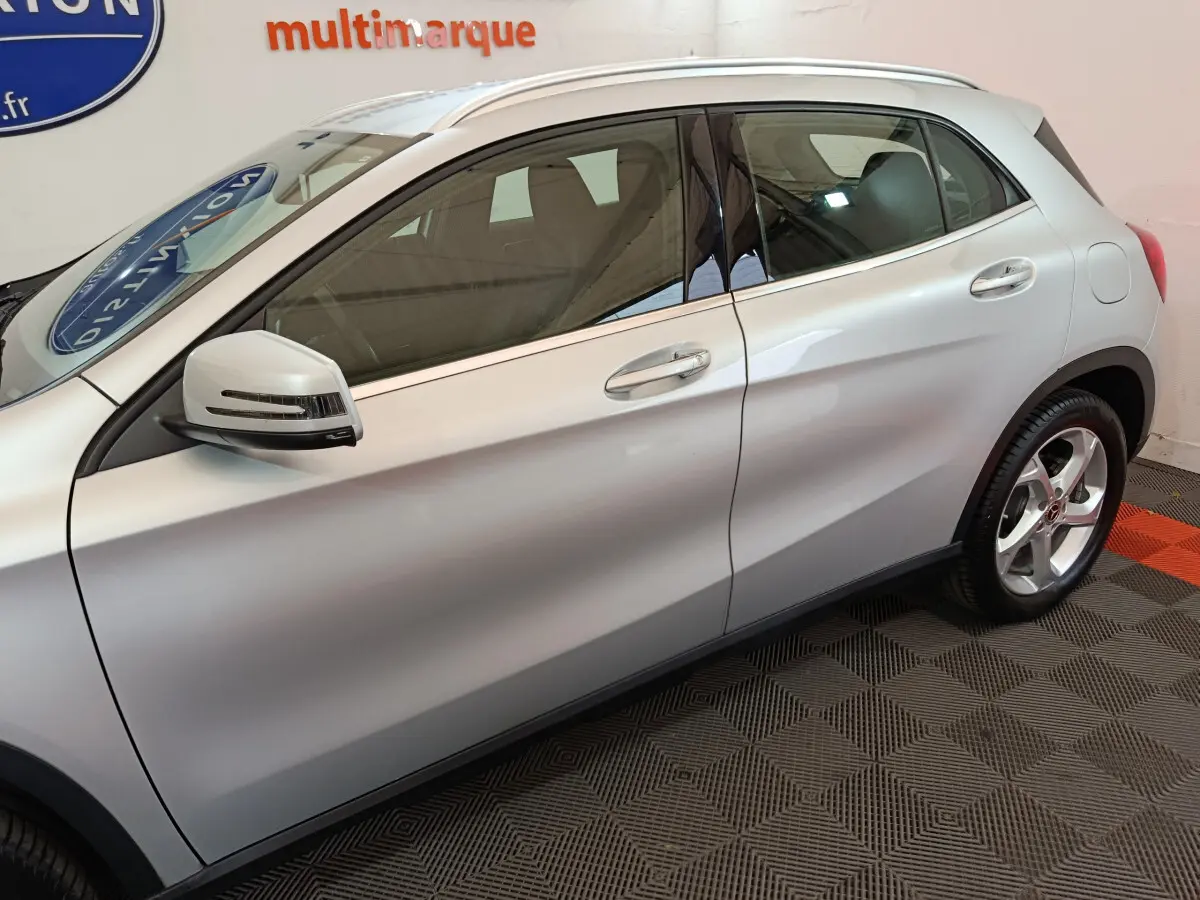 Profil côté gauche d'un Mercedes GLA 200 d gris clair avec rétroviseur et jante alliage visibles en intérieur showroom.