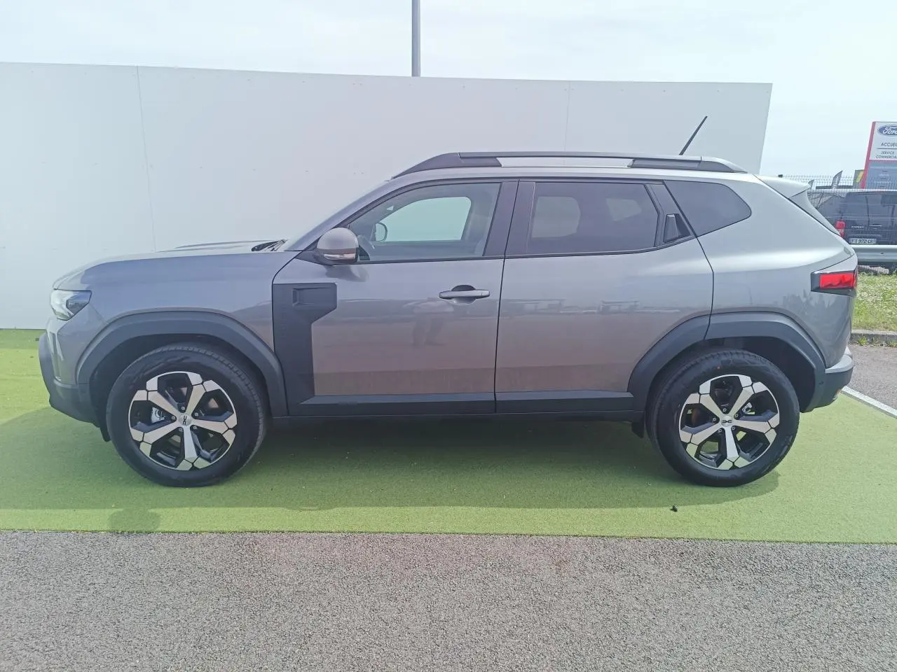 Vue de profil côté gauche du Dacia Duster III gris schiste métal avec jantes alliage 18 pouces diamantées et barres de toit noires.