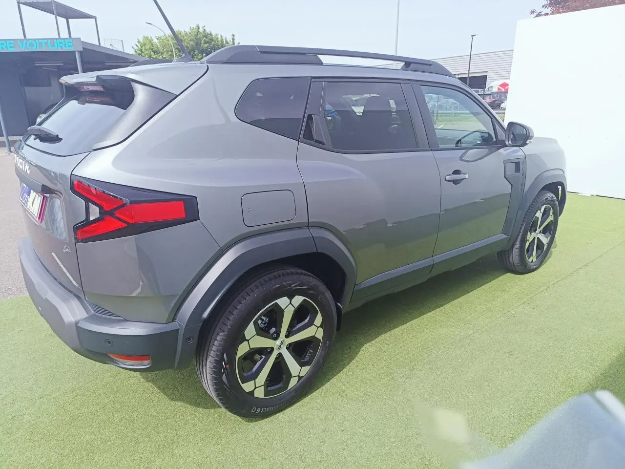 Vue 3/4 arrière droite du Dacia Duster 2026 gris schiste métal avec jantes alliage 18 pouces et barres de toit noires.