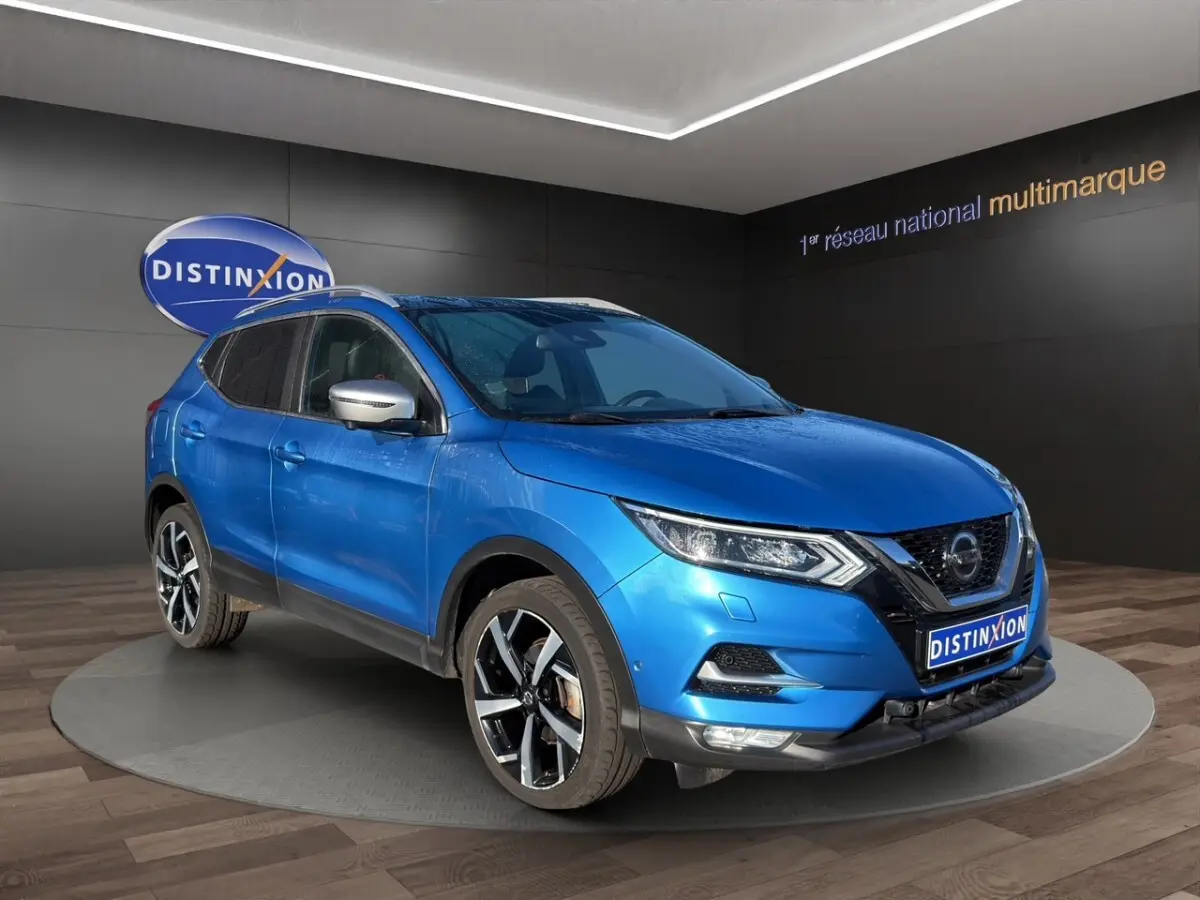 Nissan Qashqai bleu en 3/4 avant droit, avec jantes alliage noires et rétroviseurs argentés, sur podium intérieur.