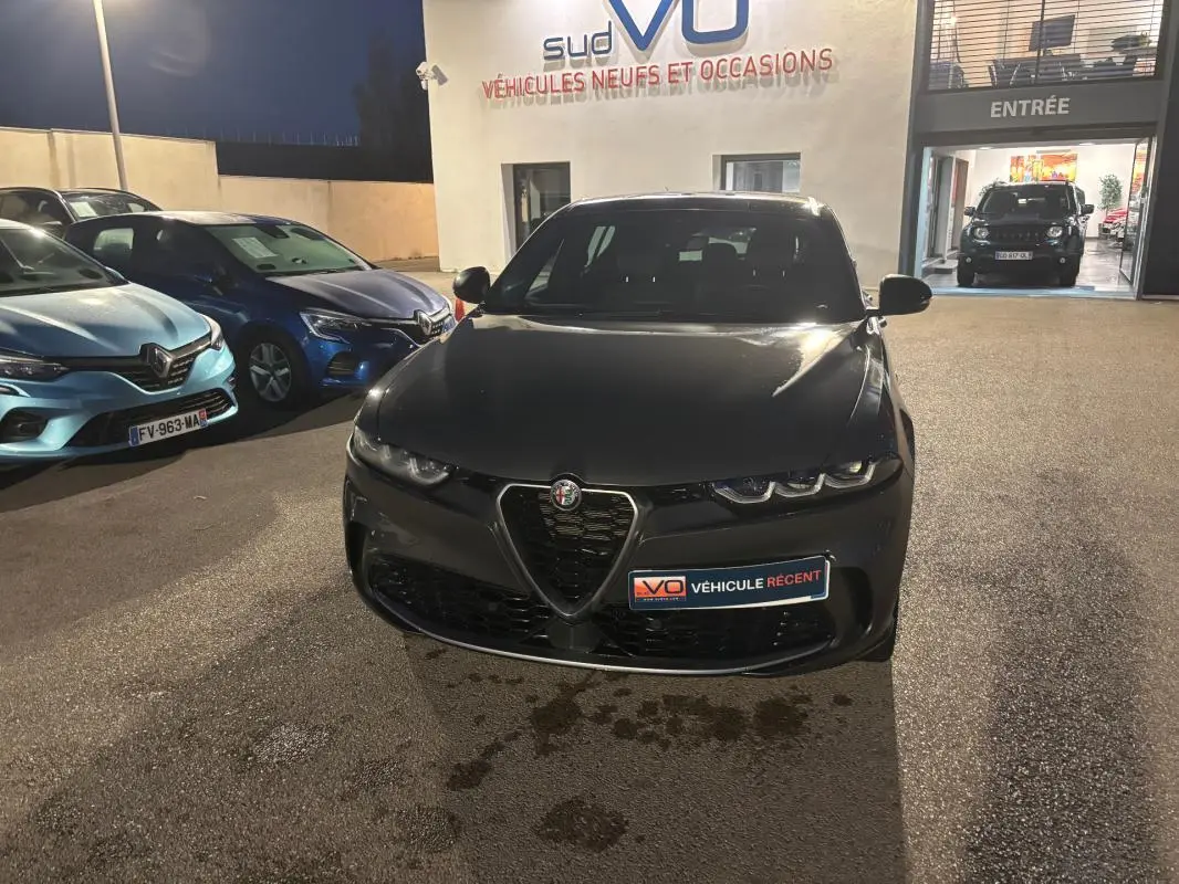 Vue avant d'une Alfa Romeo Tonale gris foncé garée de nuit devant un concessionnaire, avec phares éteints.