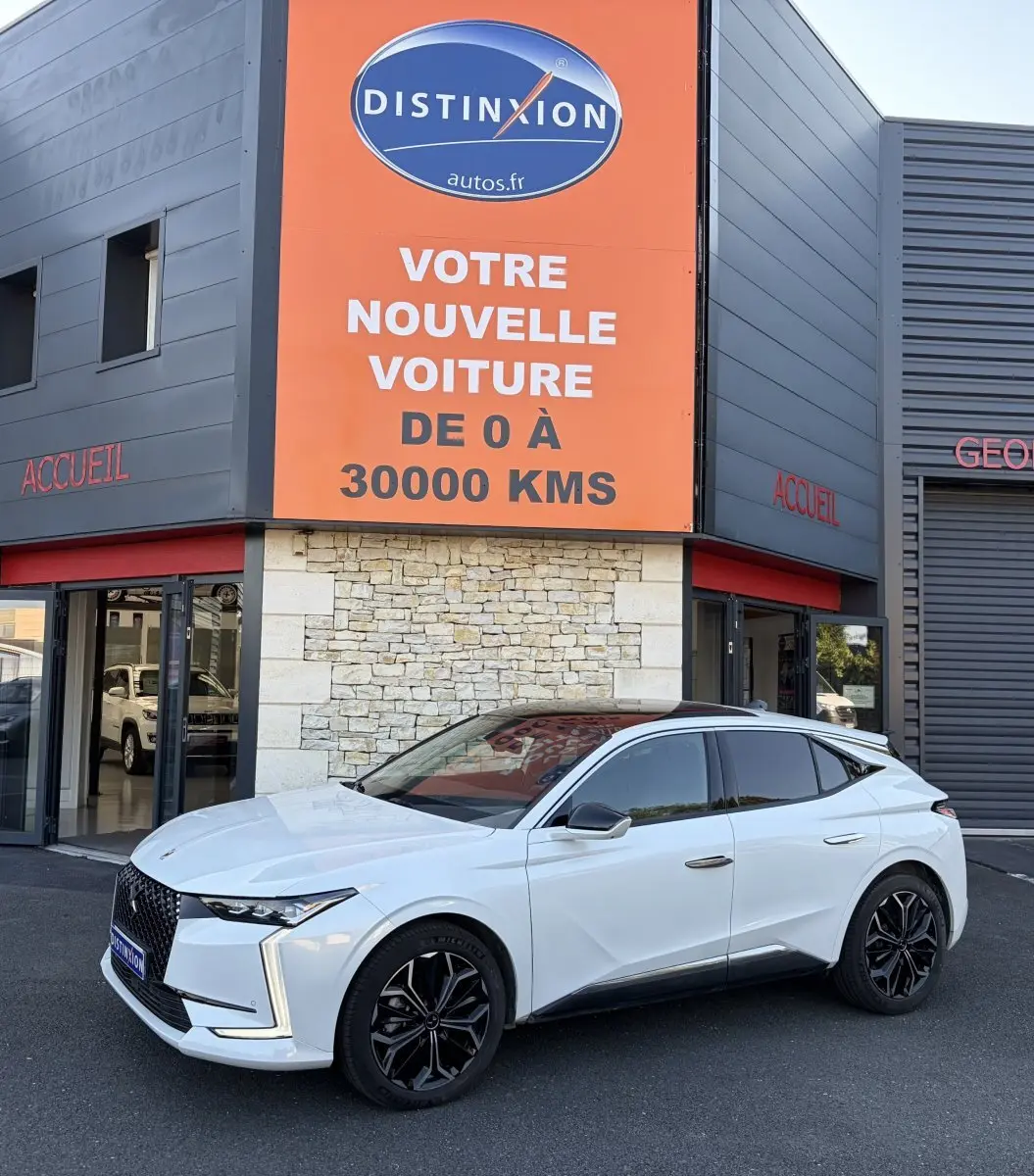 DS4 blanc vue 3/4 avant droit, jantes noires, calandre distinctive, devant un bâtiment avec panneau orange Distinxion.