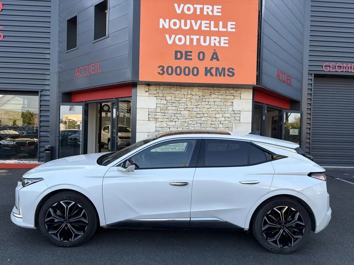 Vue latérale gauche d'une DS4 blanche 2024 avec jantes noires et vitres teintées, devant un bâtiment commercial.