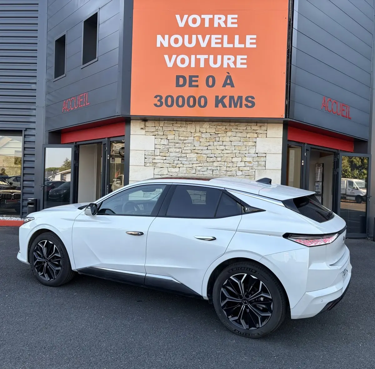 Vue côté gauche arrière d'une DS4 blanche 2024 avec jantes noires, stationnée devant un bâtiment commercial.