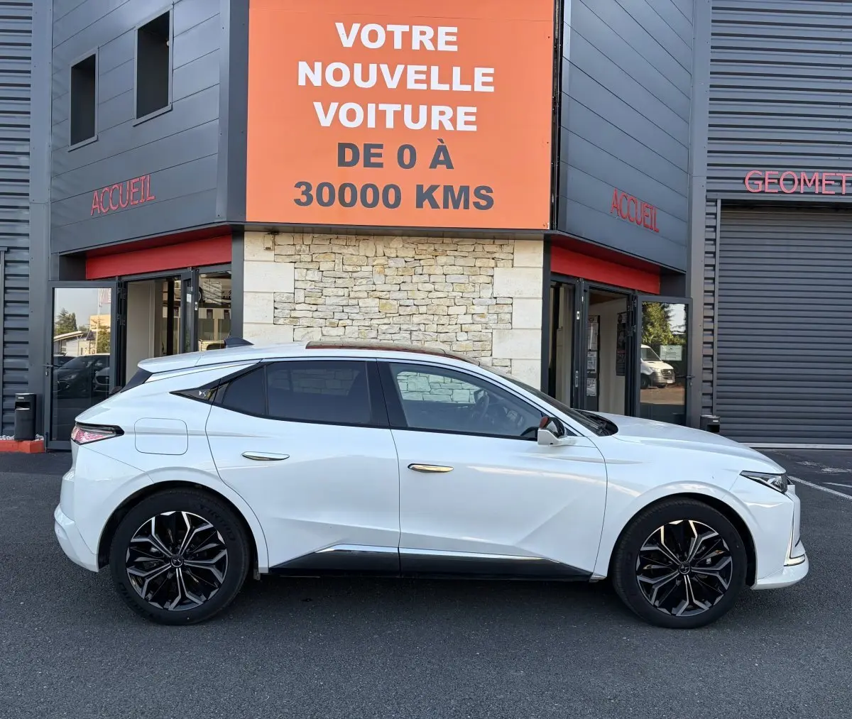 Vue de profil côté gauche d’une DS4 blanche 2024 avec jantes noires et toit panoramique, devant un bâtiment commercial.