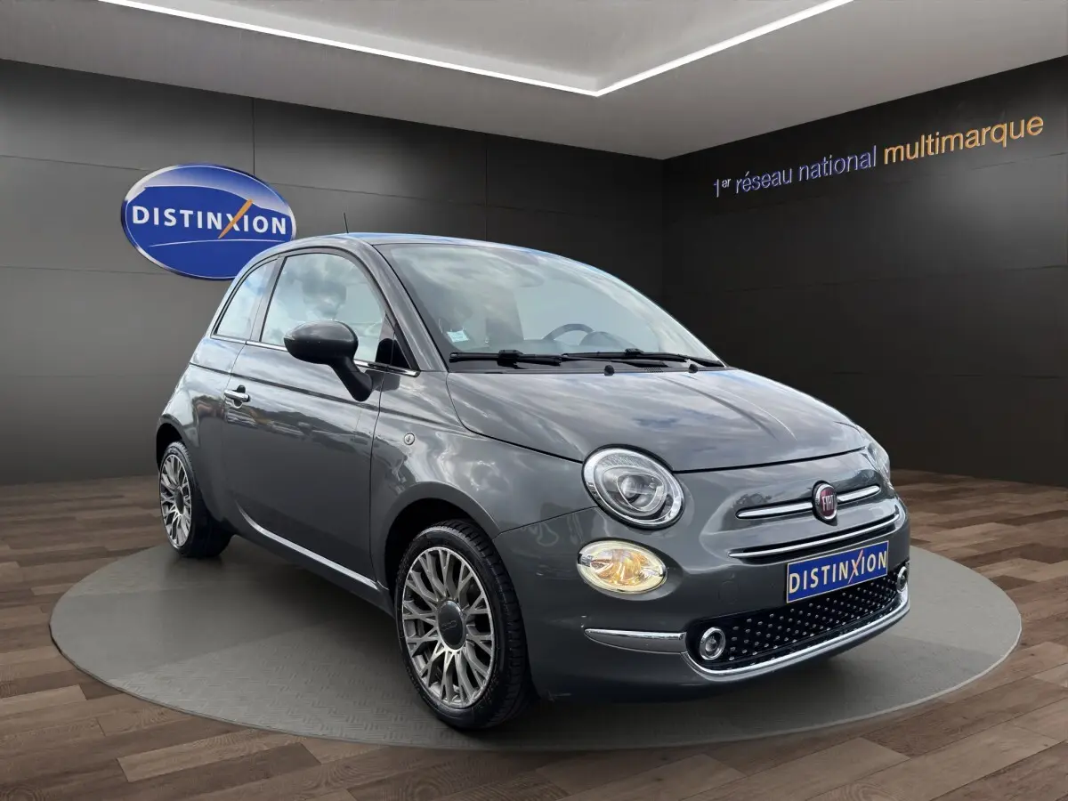 Fiat 500 gris foncé vue 3/4 avant avec phares ronds et jantes alliage détaillées sur fond showroom.