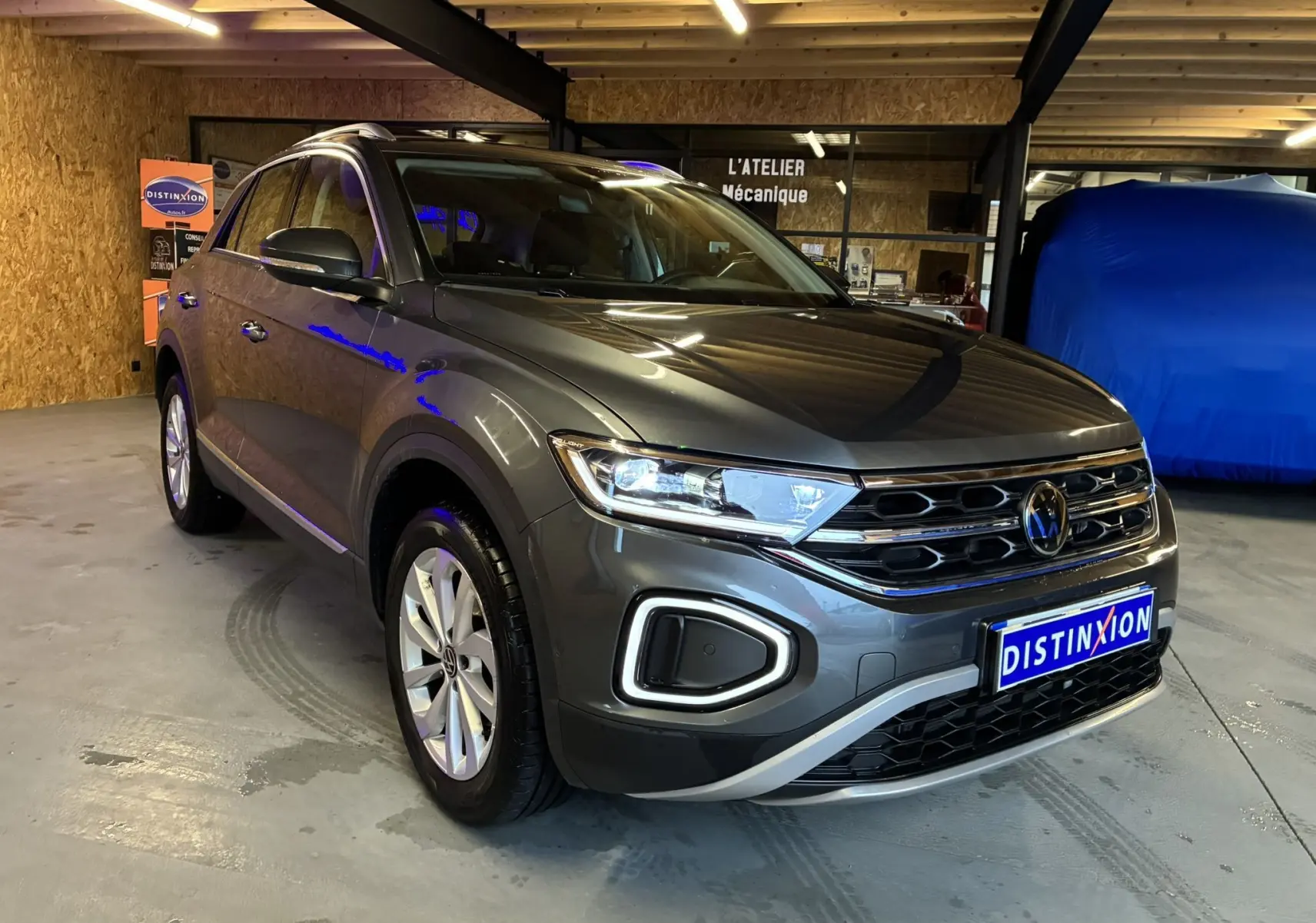 Volkswagen T-Roc gris 3/4 avant droit, phare allumé, calandre chromée et plaque Distinxion visible en intérieur d'atelier.