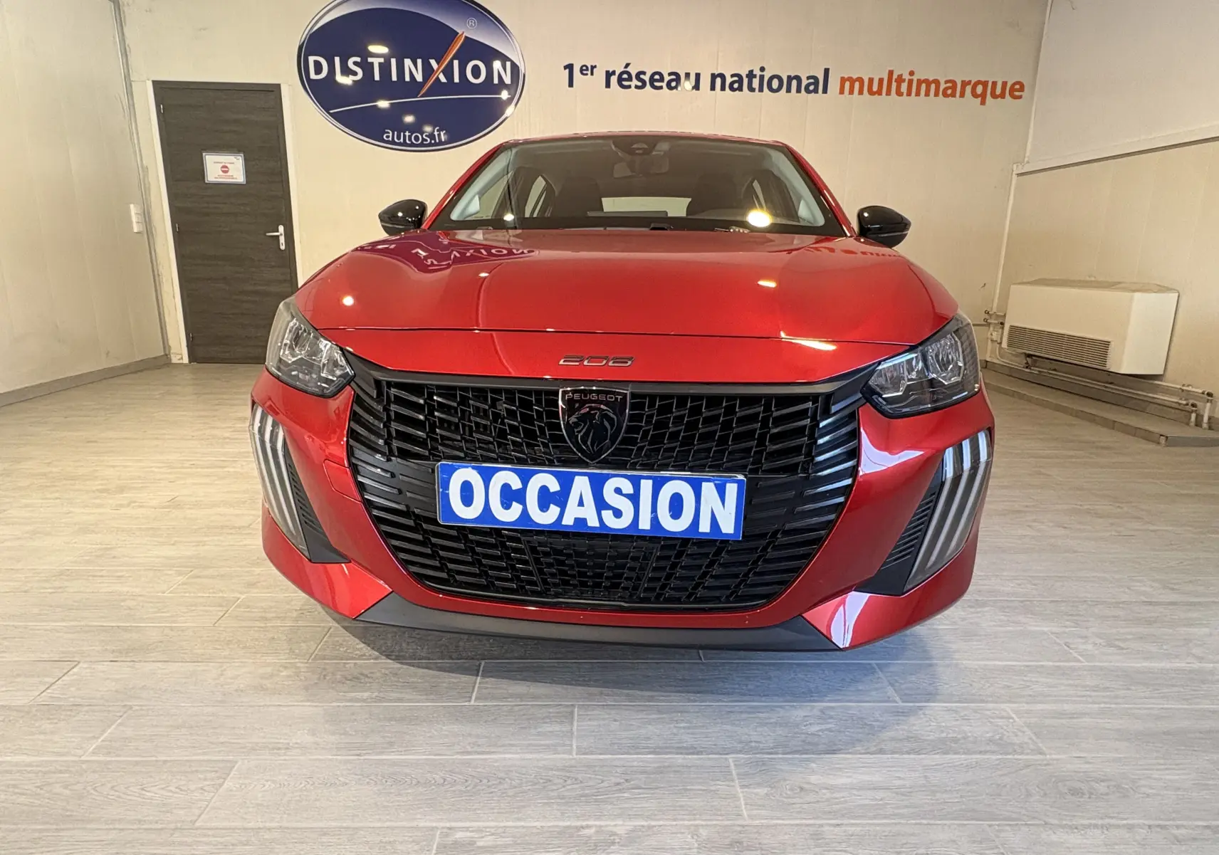 Vue frontale d'une Peugeot 208 Active rouge 2024 avec calandre noire et plaque "OCCASION" en intérieur showroom.