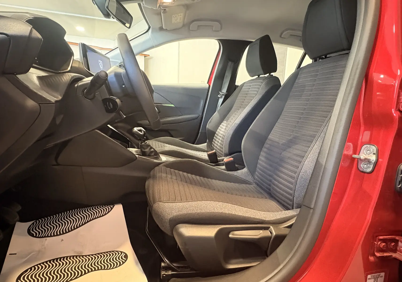 Vue intérieure côté gauche de la Peugeot 208 Active rouge 2024, montrant les sièges tissu noir et la boîte manuelle.