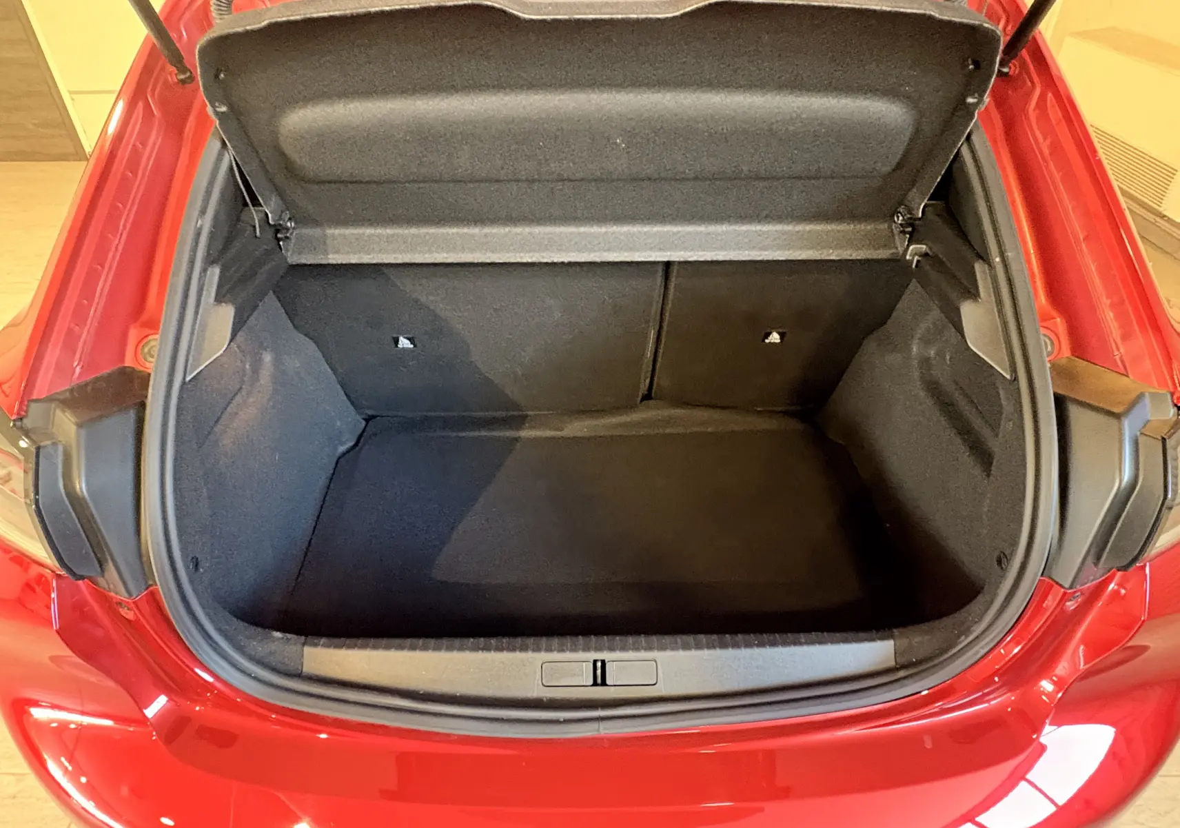 Coffre ouvert vu de dessus d'une Peugeot 208 Active rouge 2024, montrant l'espace de chargement vide et la garniture noire.