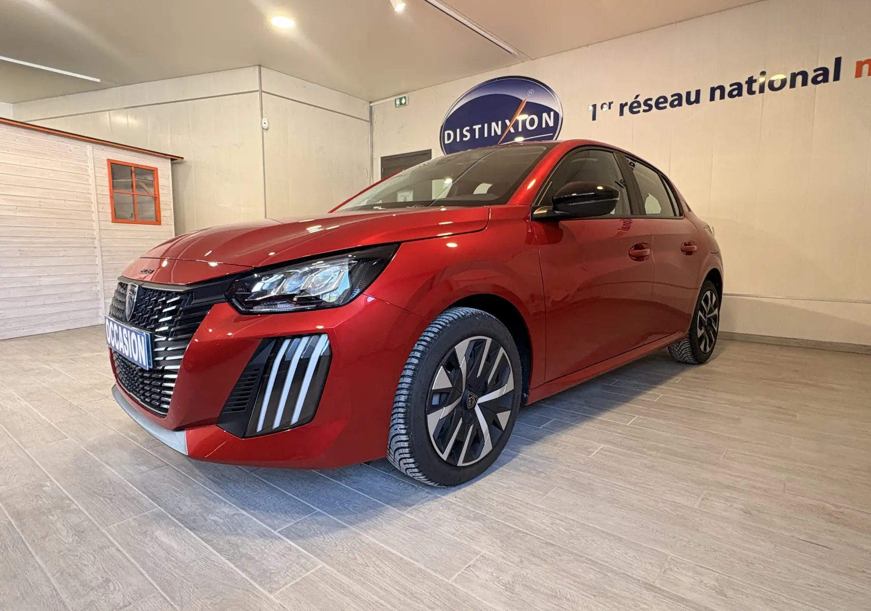 Peugeot 208 Active rouge vue 3/4 avant droit en intérieur, avec calandre noire et feux LED distinctifs.