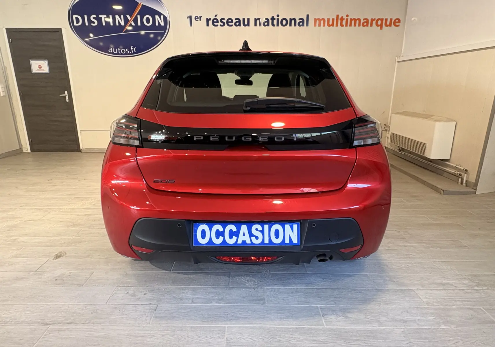 Vue arrière d'une Peugeot 208 Active rouge 2024 avec feux arrière noirs et plaque "OCCASION" en intérieur showroom.
