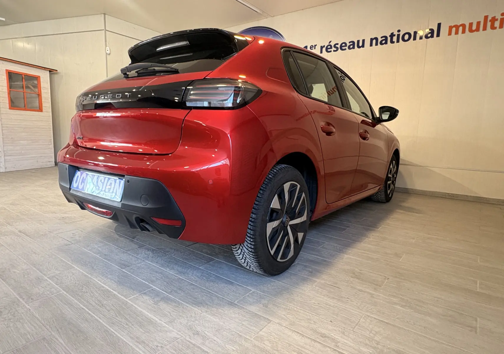 Vue 3/4 arrière droite d'une Peugeot 208 Active rouge 2024 avec feux arrière noirs et jantes alliage bicolores.