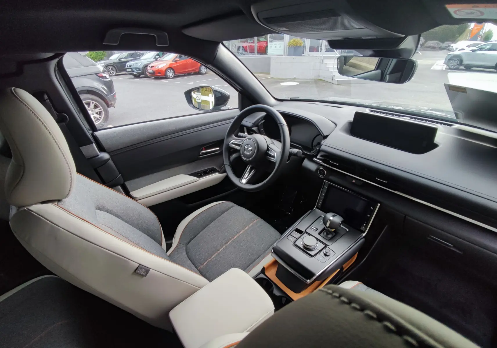 Vue intérieure côté conducteur de la Mazda MX-30 2023, sièges gris clair et foncé avec surpiqûres orange et tableau de bord moderne noir.