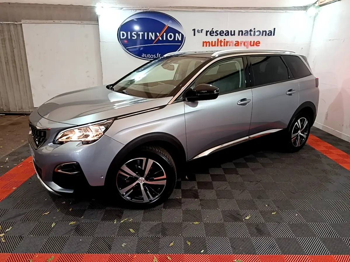 Peugeot 5008 gris clair vue 3/4 avant droit, avec jantes alliage et barres de toit chromées en intérieur showroom.