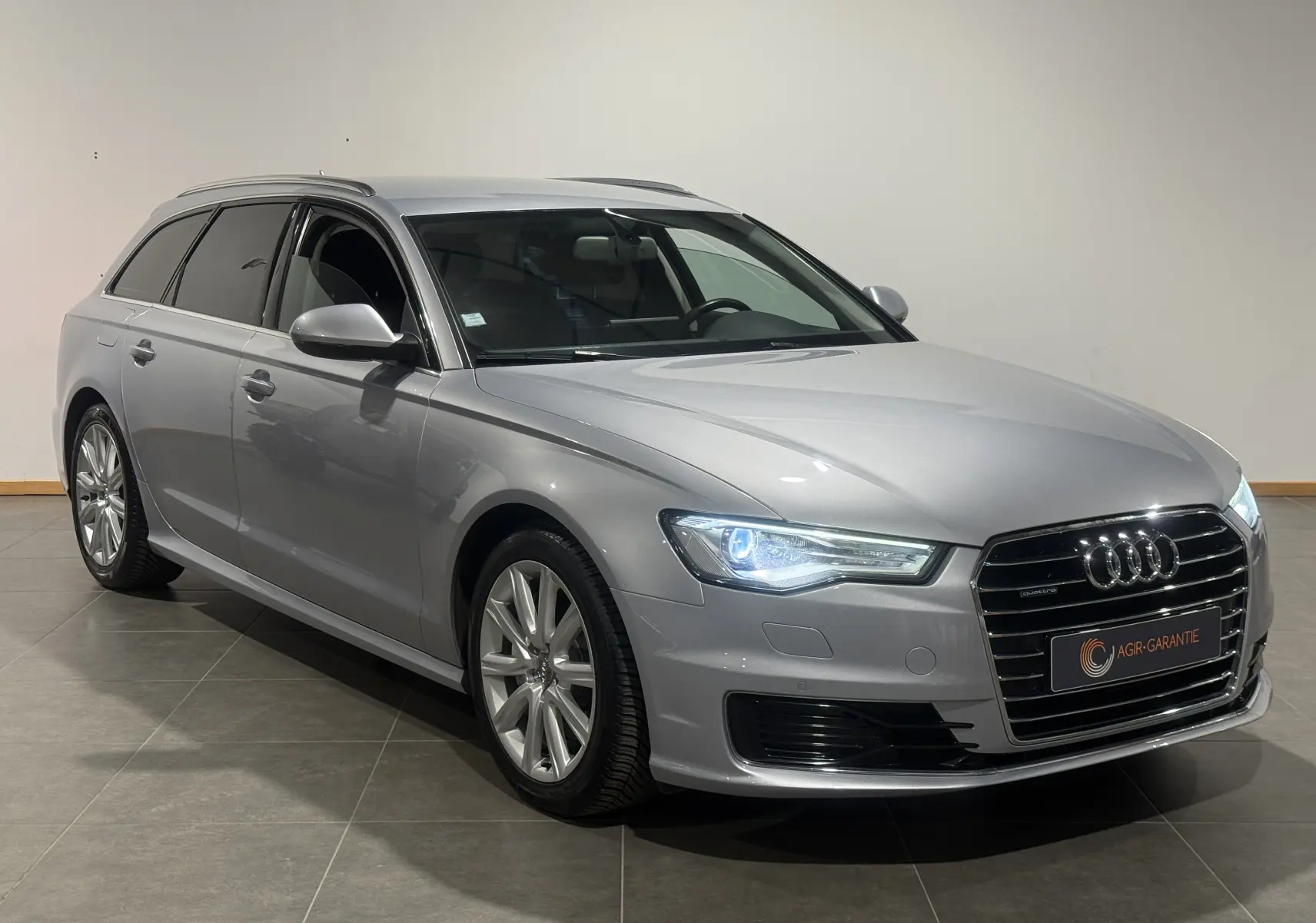 Audi A6 Avant gris en vue 3/4 avant droit, phares allumés et calandre chromée distinctive.