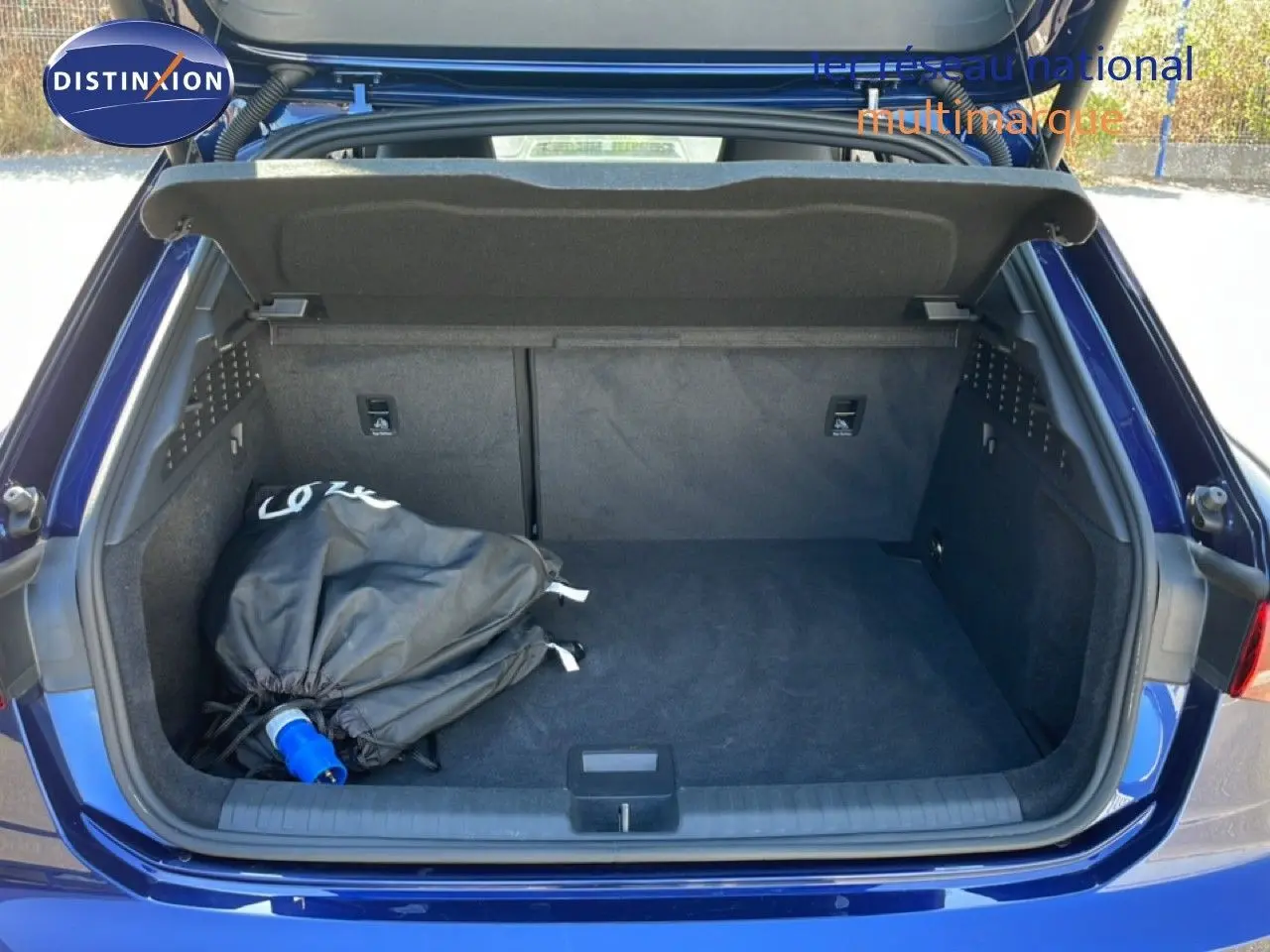Coffre ouvert vu de l'arrière d'une Audi A3 Sportback bleu Navarre, avec un sac noir et prise de recharge visible.