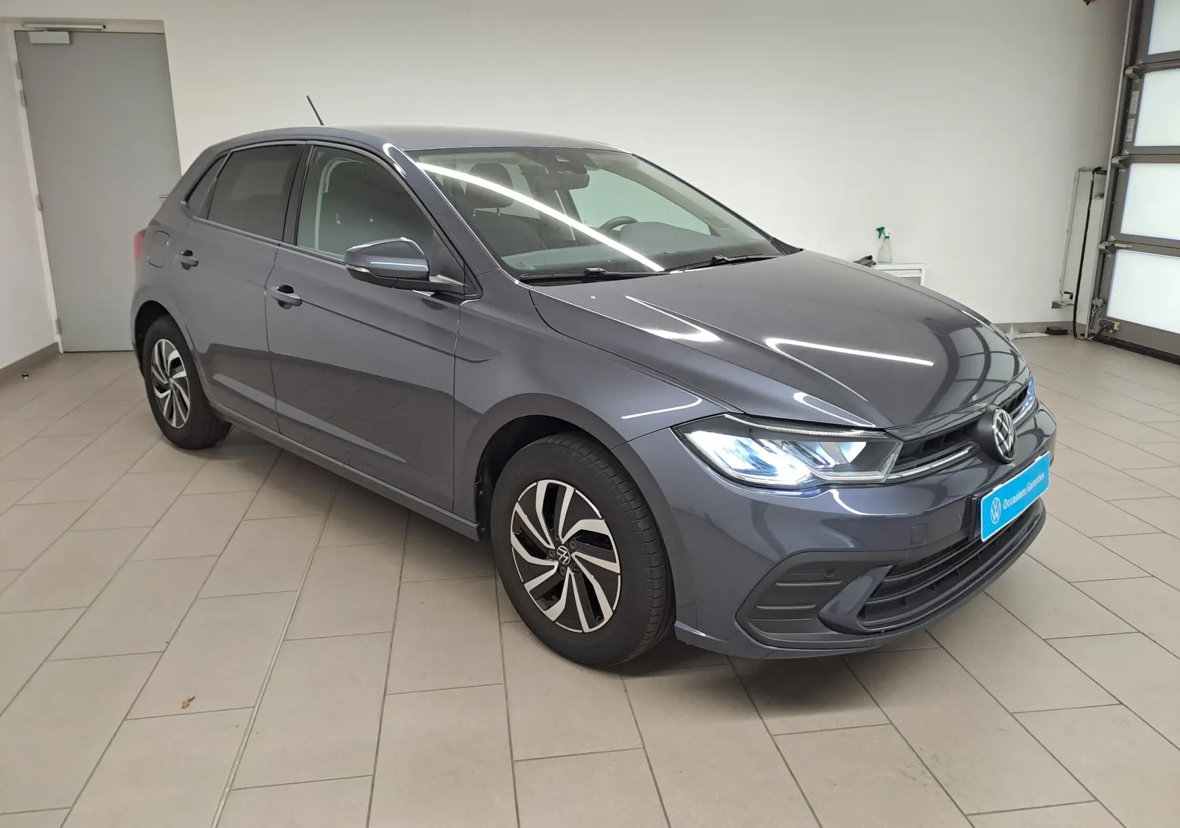 Volkswagen Polo gris cendre vue 3/4 avant droit en intérieur avec phares allumés et jantes distinctives.