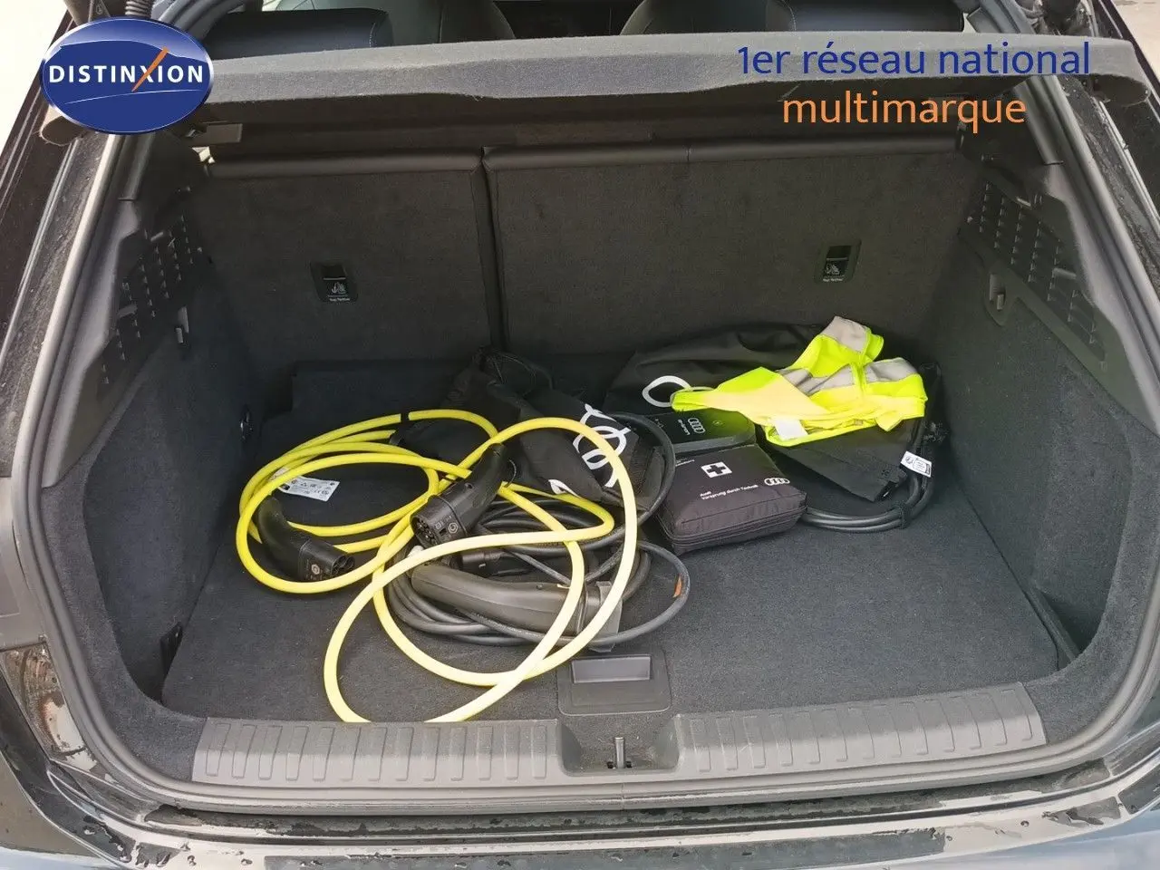 Coffre ouvert vu de l'arrière d'une Audi A3 Sportback noir mito metal avec câbles de recharge et gilet jaune.