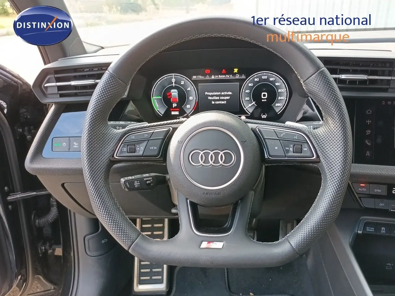 Vue intérieure centrée sur le volant perforé d’une Audi A3 Sportback noire, avec tableau de bord numérique et commandes intégrées.