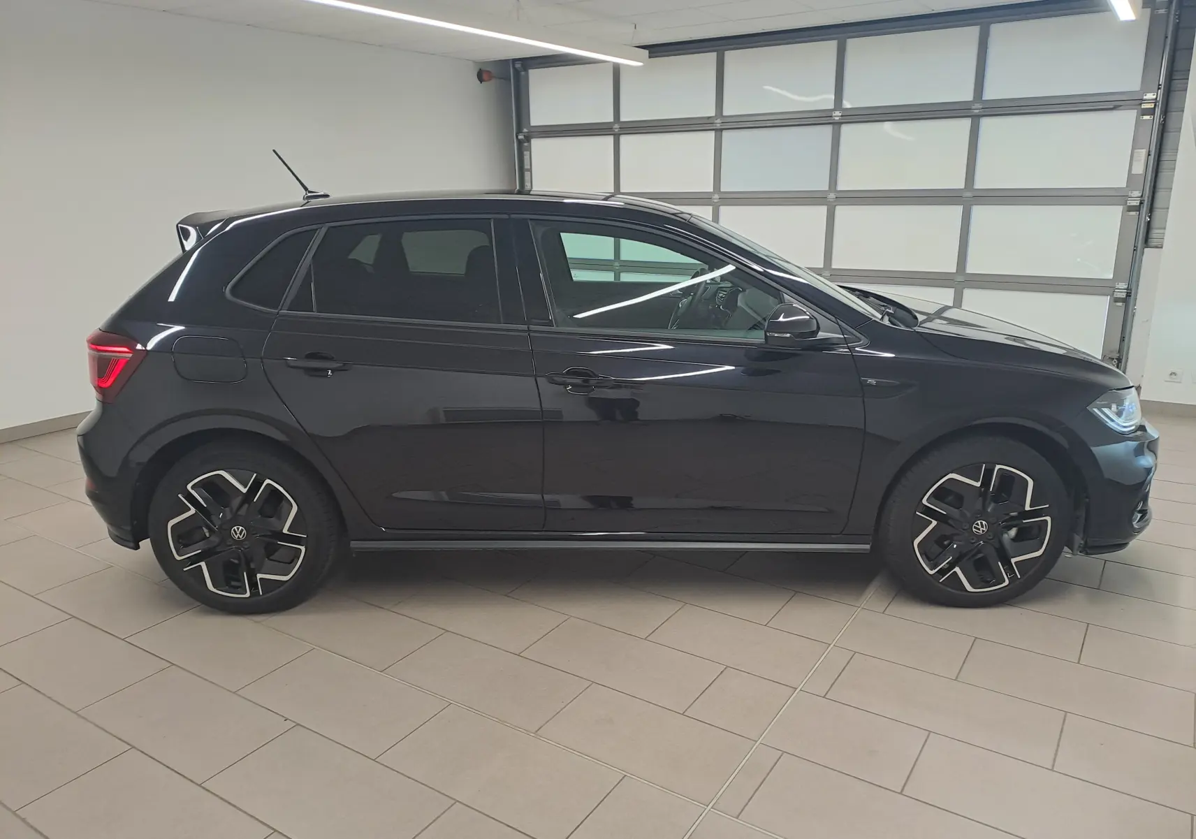 Vue de profil côté gauche d'une Volkswagen Polo 2025 noire intense avec jantes R-Line distinctives en intérieur lumineux.