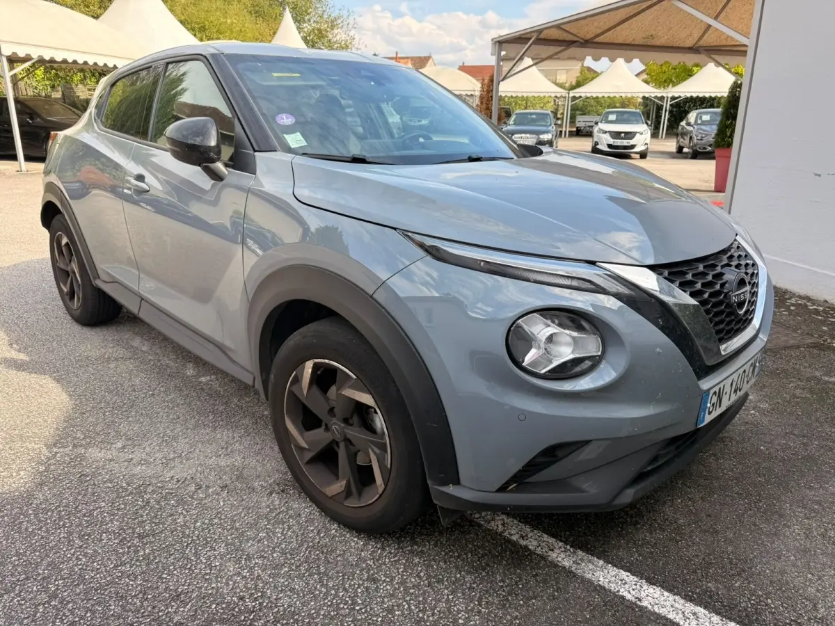 Nissan Juke gris clair vu en 3/4 avant droit, avec jantes noires et calandre distinctive en V chromé.
