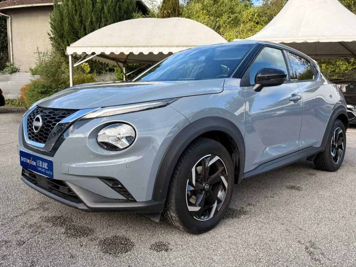 Nissan Juke gris clair vu de 3/4 avant droit, avec phares ronds et jantes noires à motifs géométriques.