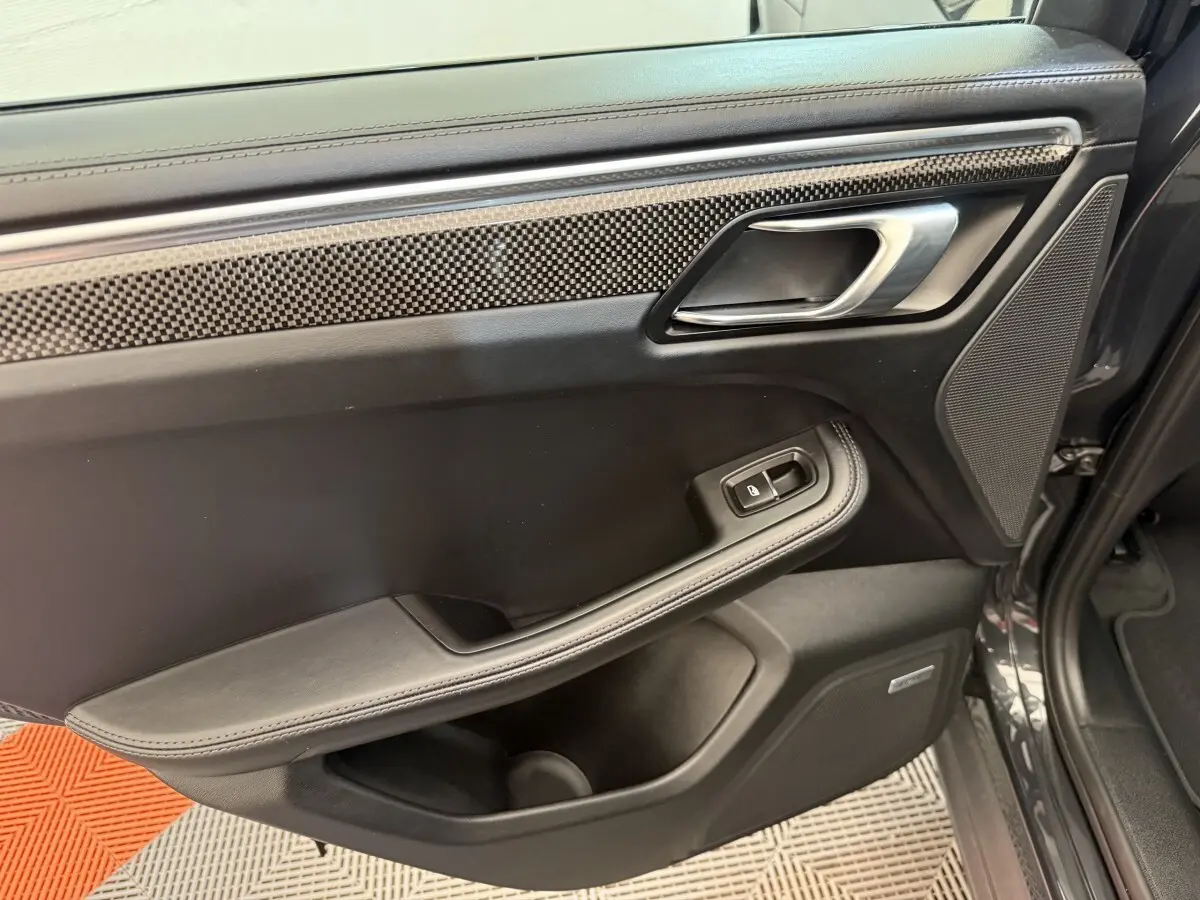 Vue intérieure côté gauche de la porte arrière d’un Porsche Macan 3.0 S gris foncé avec garniture carbone et bouton de lève-vitre.