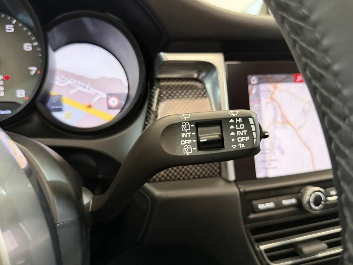 Gros plan sur la commande d’essuie-glace à gauche du volant en cuir du Porsche Macan 3.0 S gris foncé, avec écran tactile en arrière-plan.