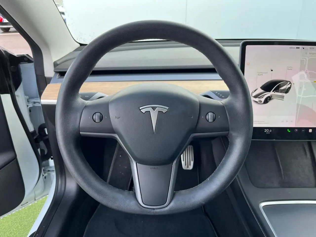 Vue centrée sur le volant noir d'une Tesla Model 3 blanche, avec écran tactile et intérieur partiellement en bois clair.