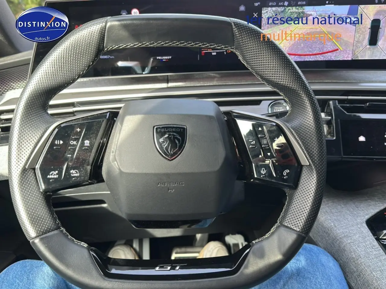 Vue rapprochée du volant cuir perforé noir avec logo Peugeot et inscription GT, tableau de bord numérique en arrière-plan.