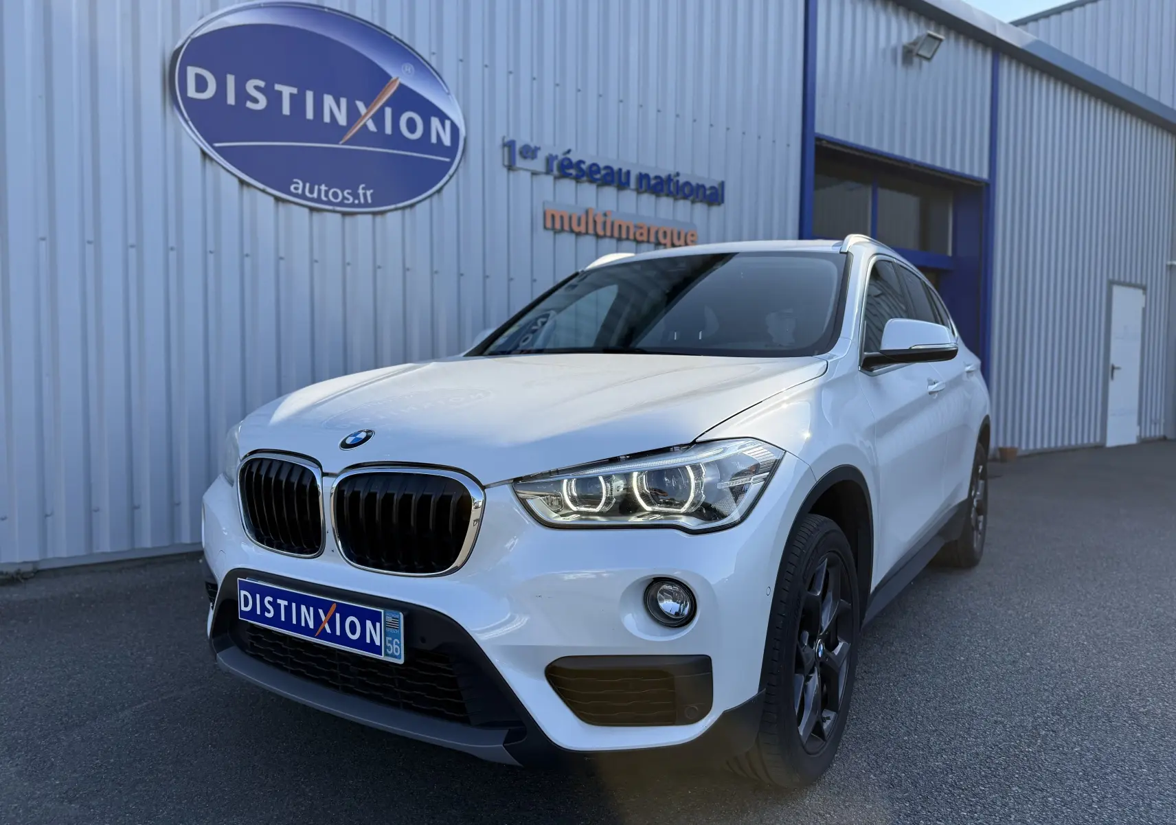 BMW X1 blanc vue 3/4 avant droit avec phares LED allumés devant un bâtiment industriel.