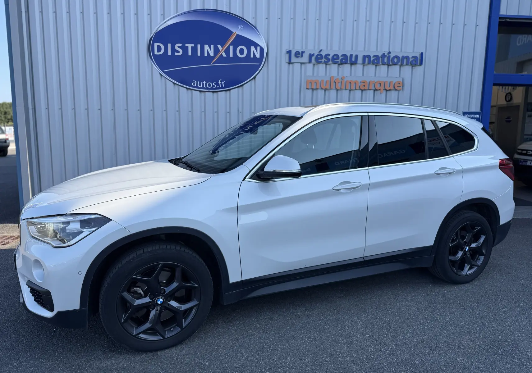 BMW X1 20D blanc vue de profil côté gauche, avec jantes noires et vitres teintées, stationné devant un bâtiment.