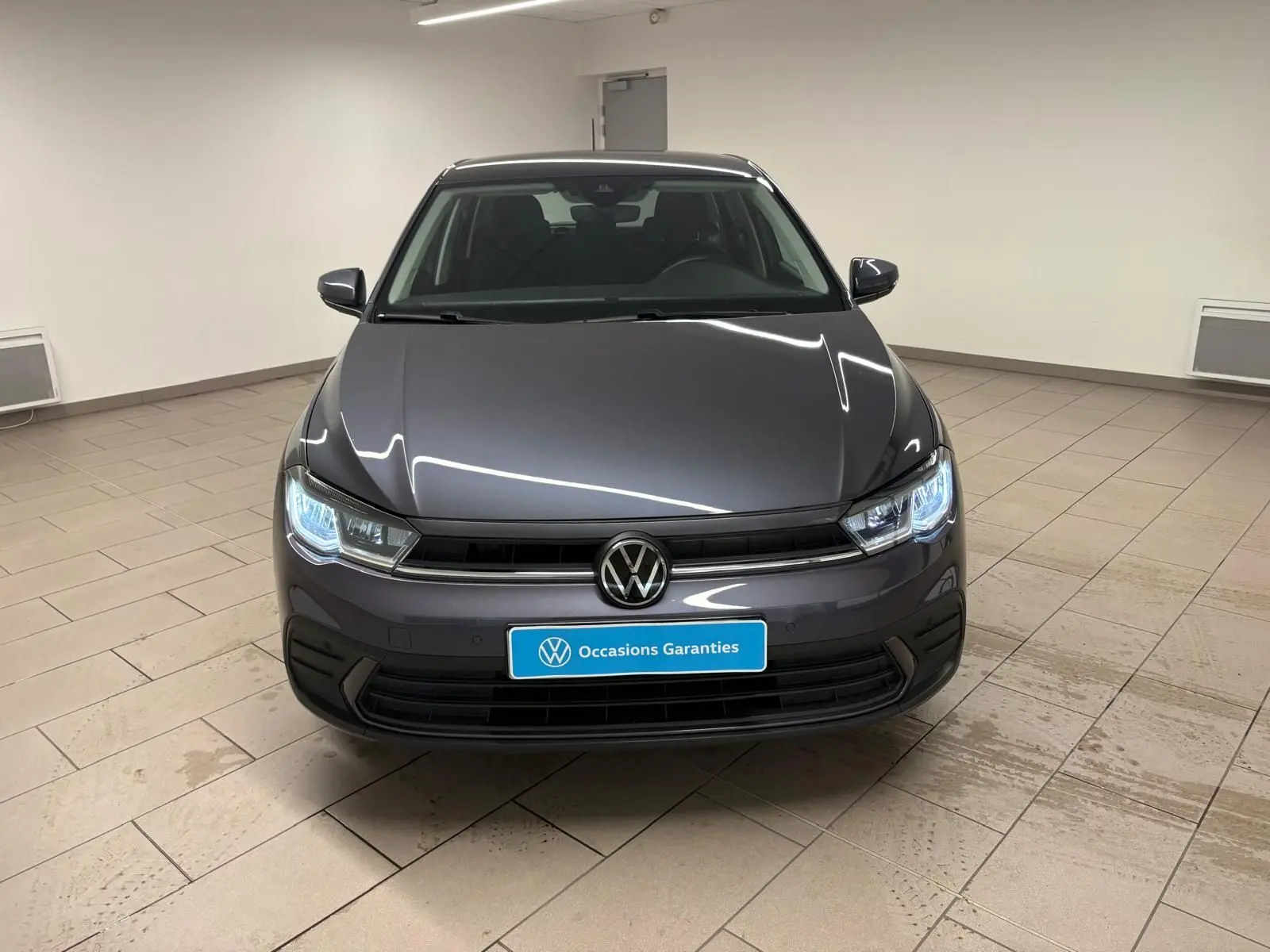 Volkswagen Polo gris cendre vue de face en intérieur, phares allumés et logo VW bien visible.