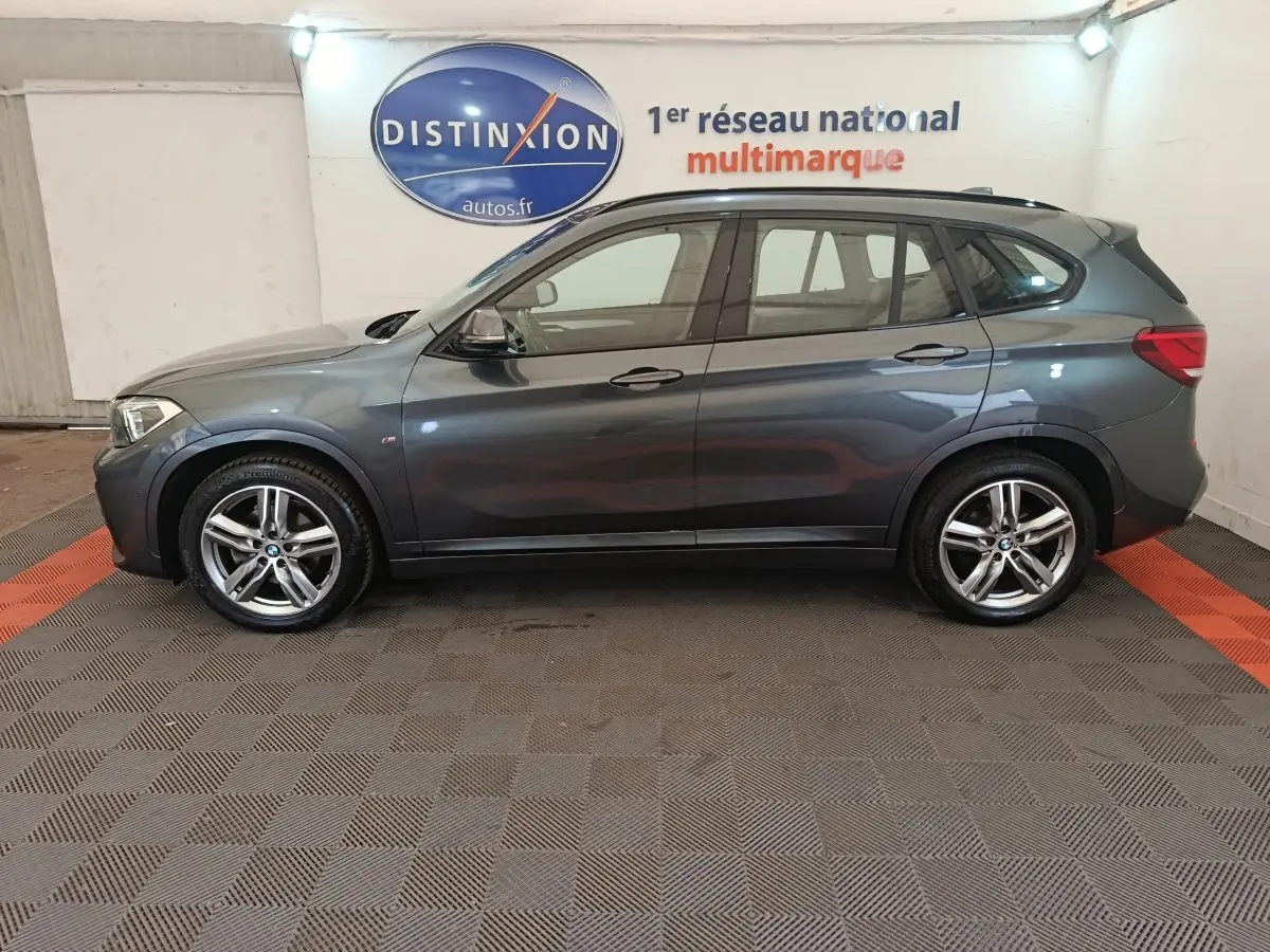 BMW X1 sDrive18i M Sport gris, vue profil côté gauche, avec jantes alliage et toit noir en intérieur showroom.
