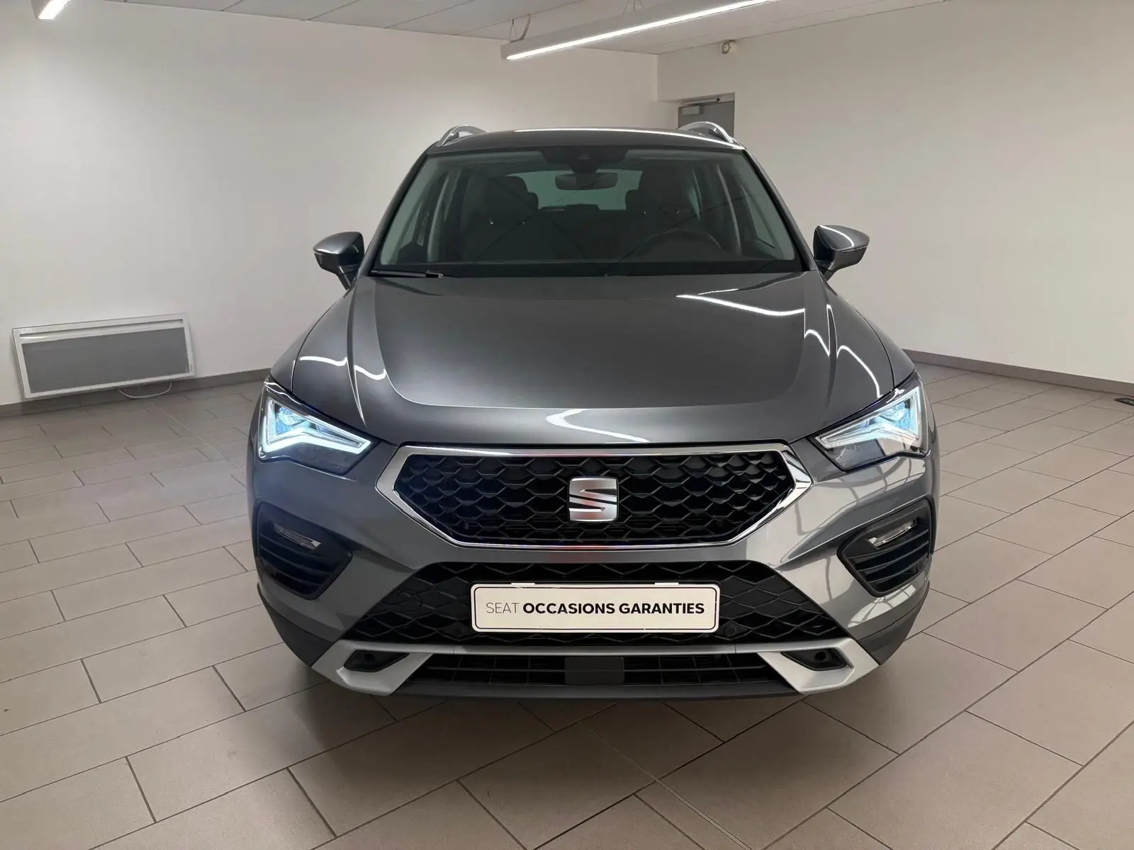 Vue frontale d'un SEAT Ateca gris graphite 2025 avec calandre hexagonale et phares LED allumés dans un showroom.