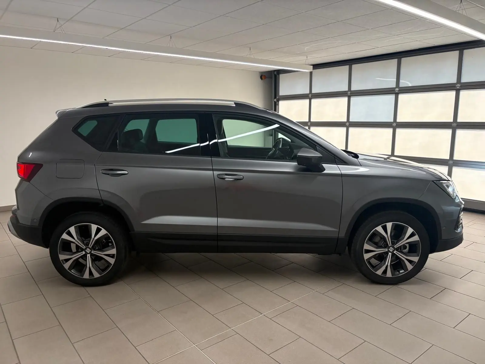 Vue de profil droit du SEAT Ateca 2025 gris graphite avec jantes alliage 18 pouces et toit noir en intérieur showroom.
