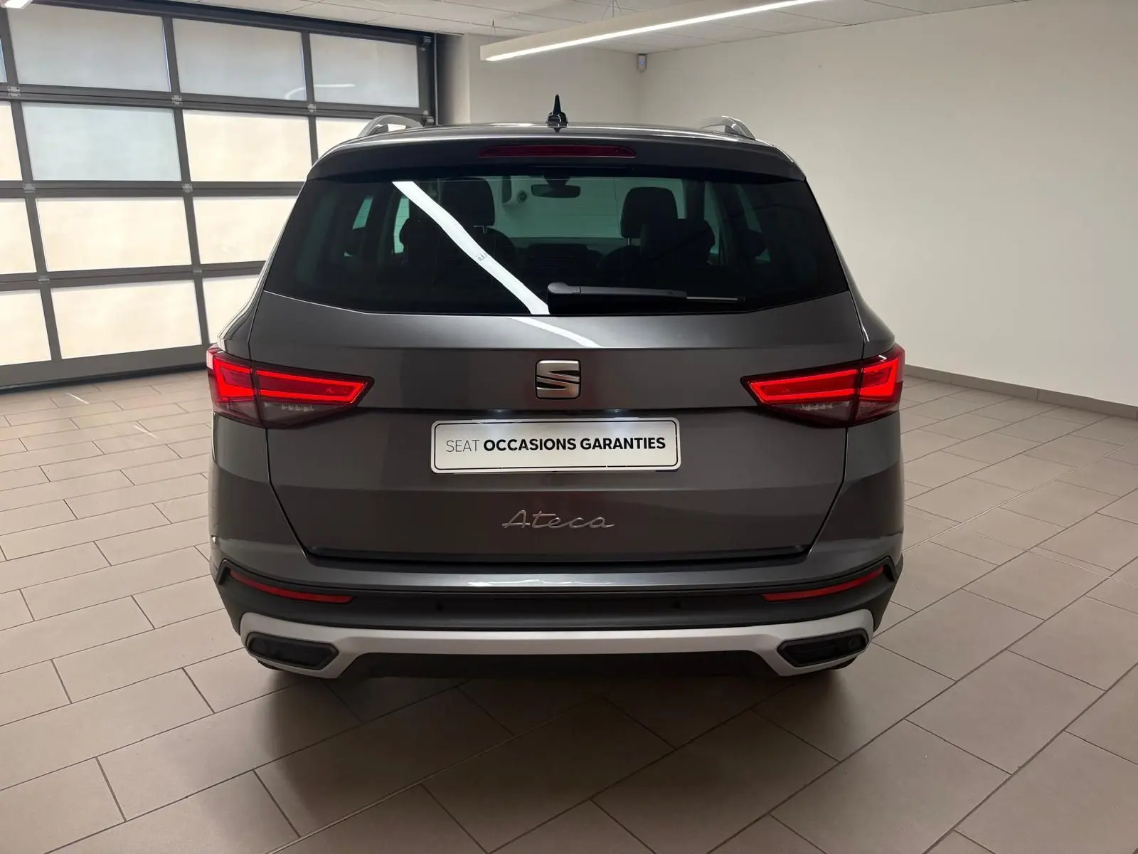 Vue arrière d'un SEAT Ateca 2.0 TDI gris graphite avec feux allumés et hayon électrique en intérieur showroom.