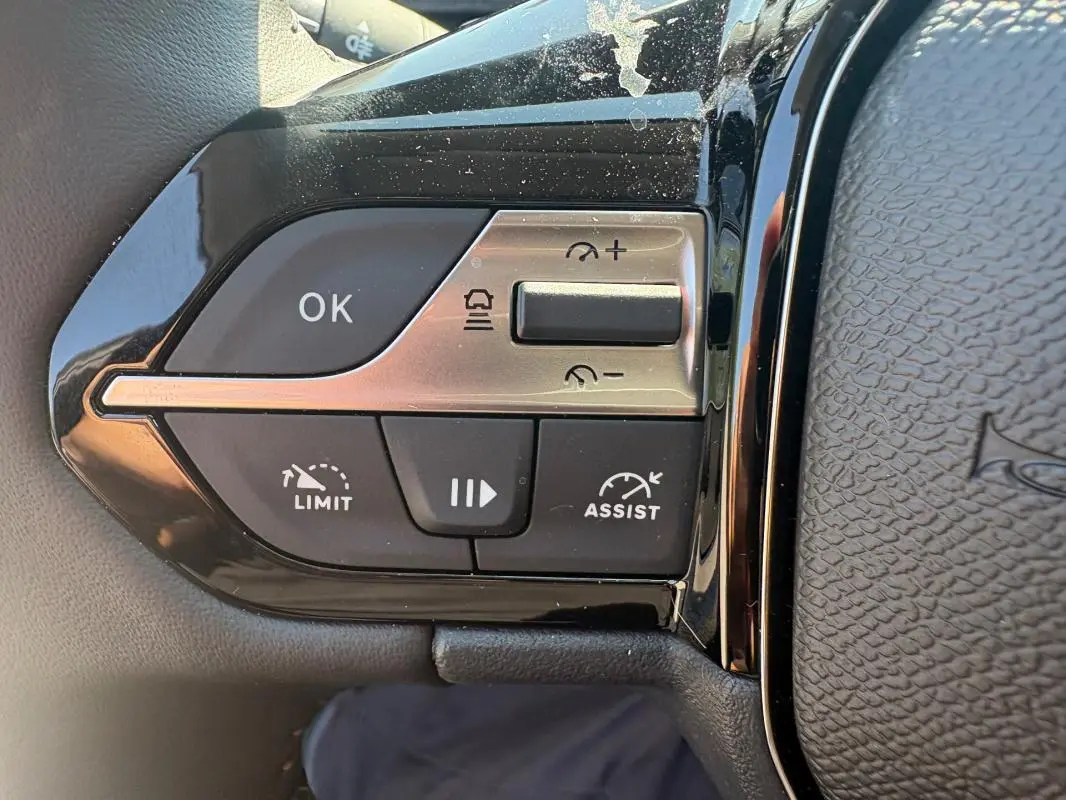 Gros plan sur les commandes de régulateur et limiteur de vitesse à gauche du volant d’une Peugeot 308 PHEV 2024 gris clair.