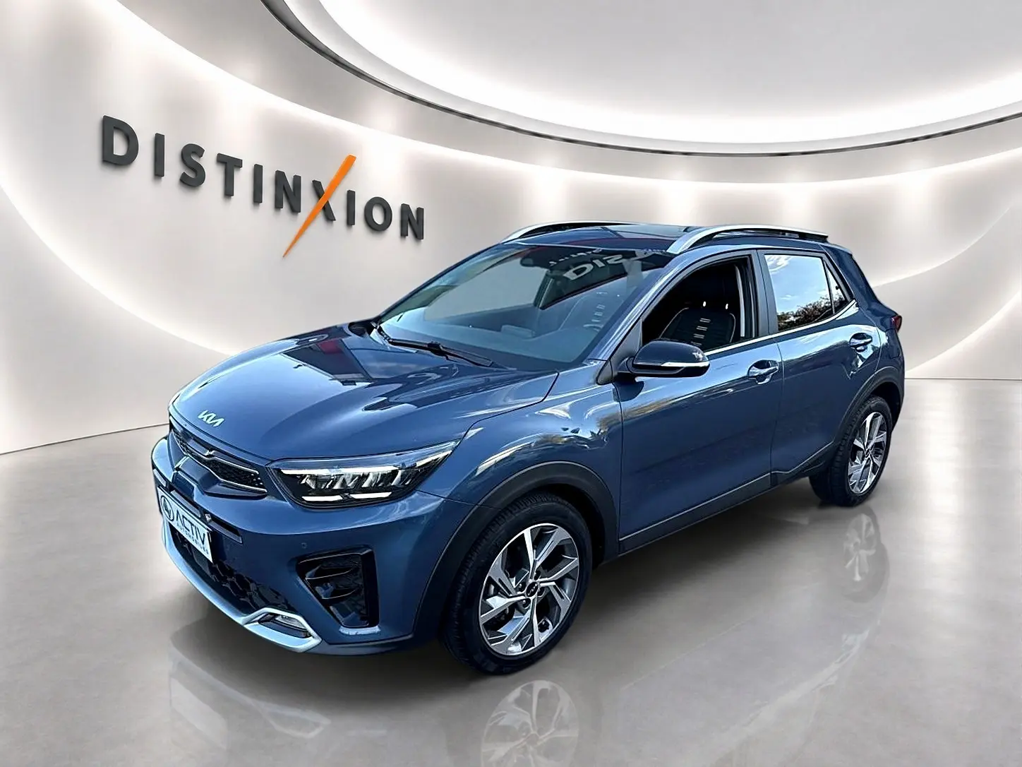 KIA Stonic gris en vue 3/4 avant droit, avec jantes alu et barres de toit visibles en showroom lumineux.