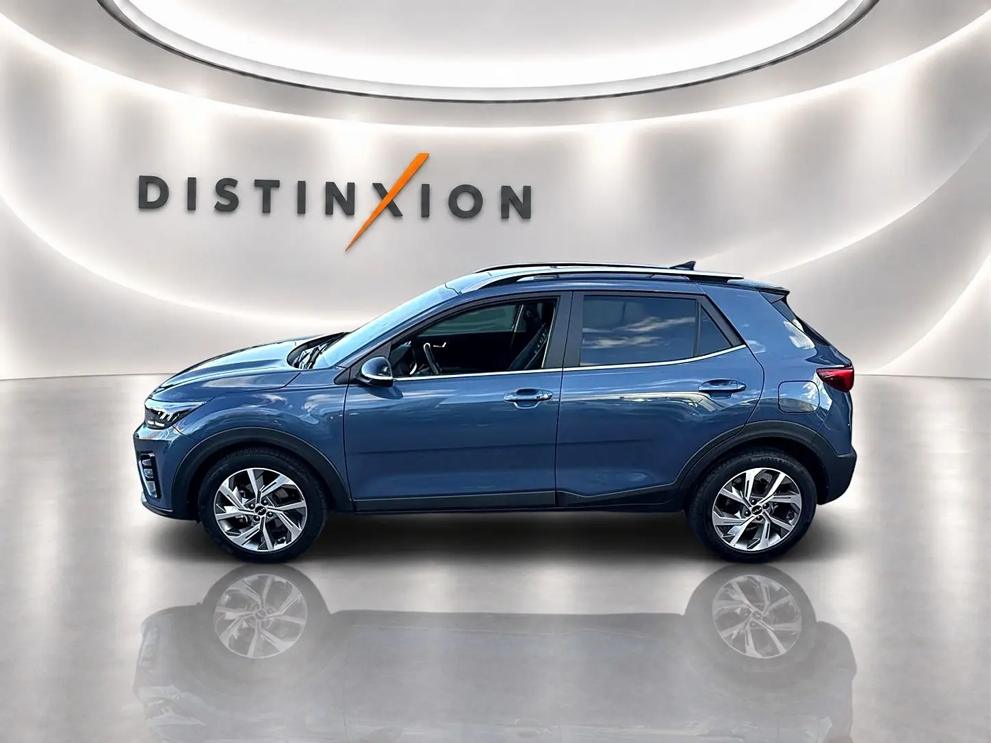 Vue de profil côté gauche d'un KIA Stonic gris 2025 dans un showroom avec jantes alliage et barres de toit.