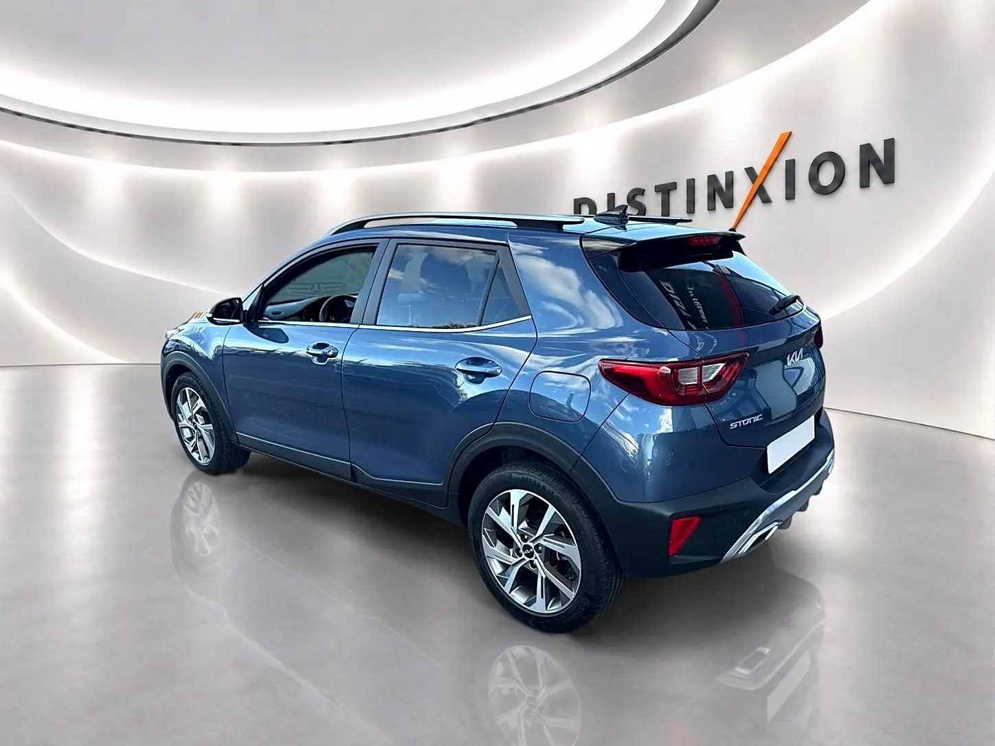 KIA Stonic gris en 3/4 arrière droit, avec barres de toit et jantes alliage visibles dans un showroom lumineux.