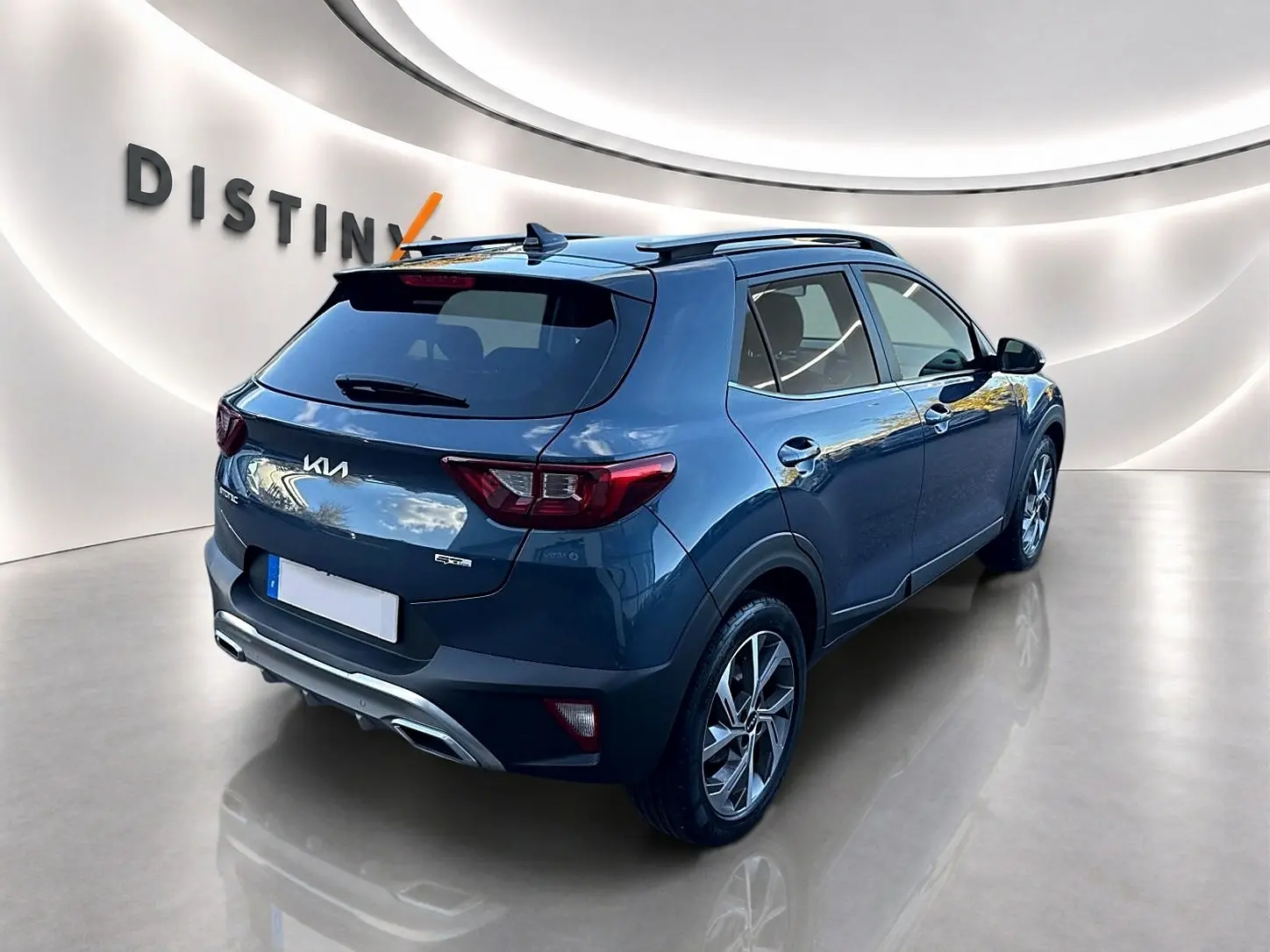 Vue 3/4 arrière droite d’un KIA Stonic gris avec jantes alu et barres de toit dans un showroom lumineux.
