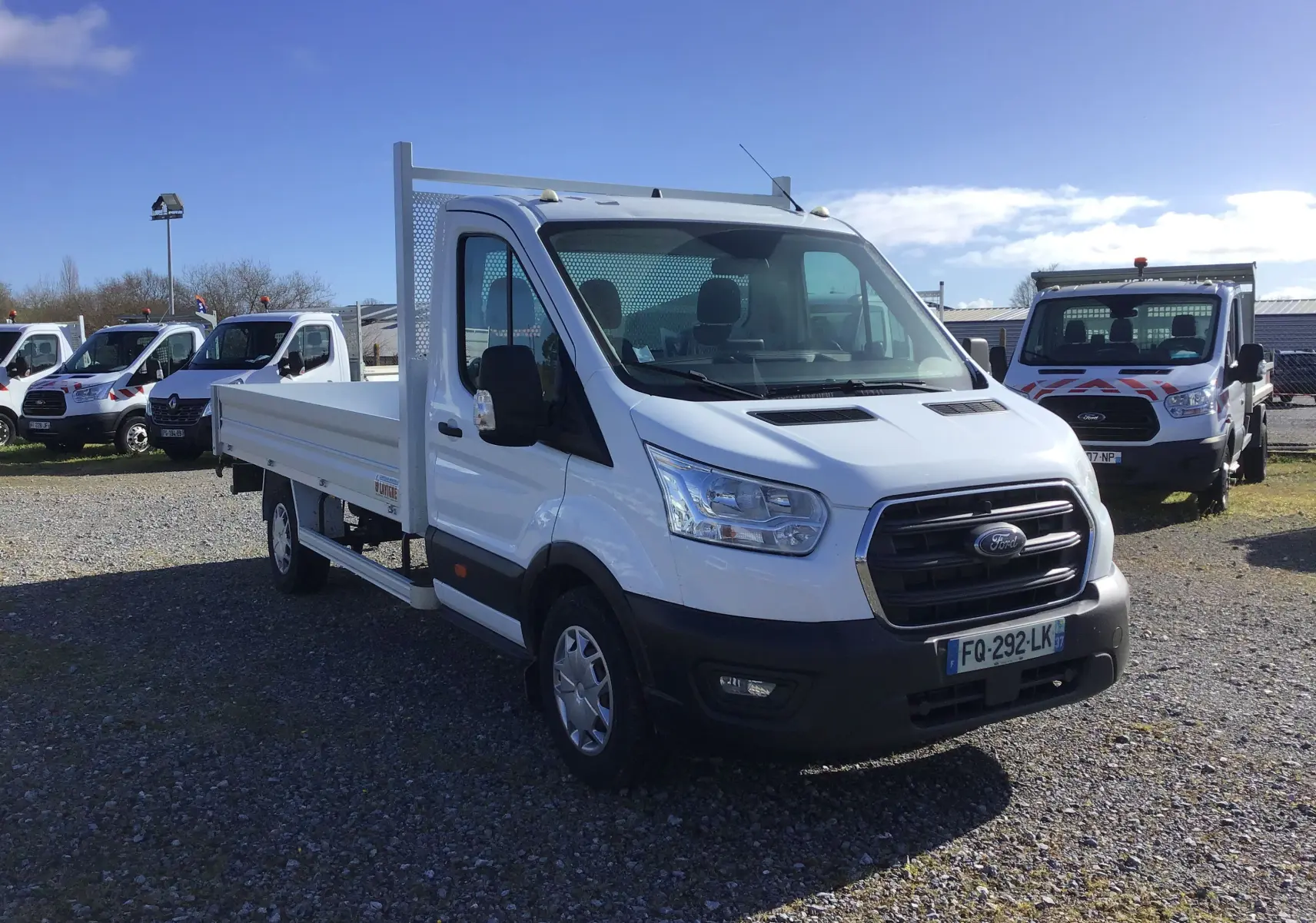 Ford Transit Chassis Cabine blanc en 3/4 avant droit, avec plateau 4 mètres et ridelles en aluminium visibles.