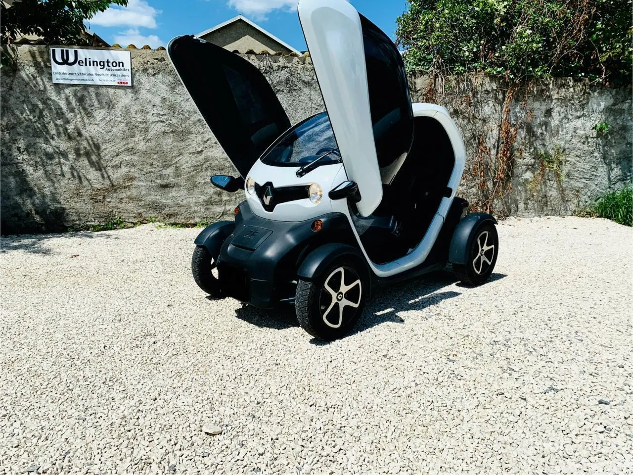 Renault Twizy blanc en 3/4 avant droit avec portes papillon ouvertes sur sol gravillonné.