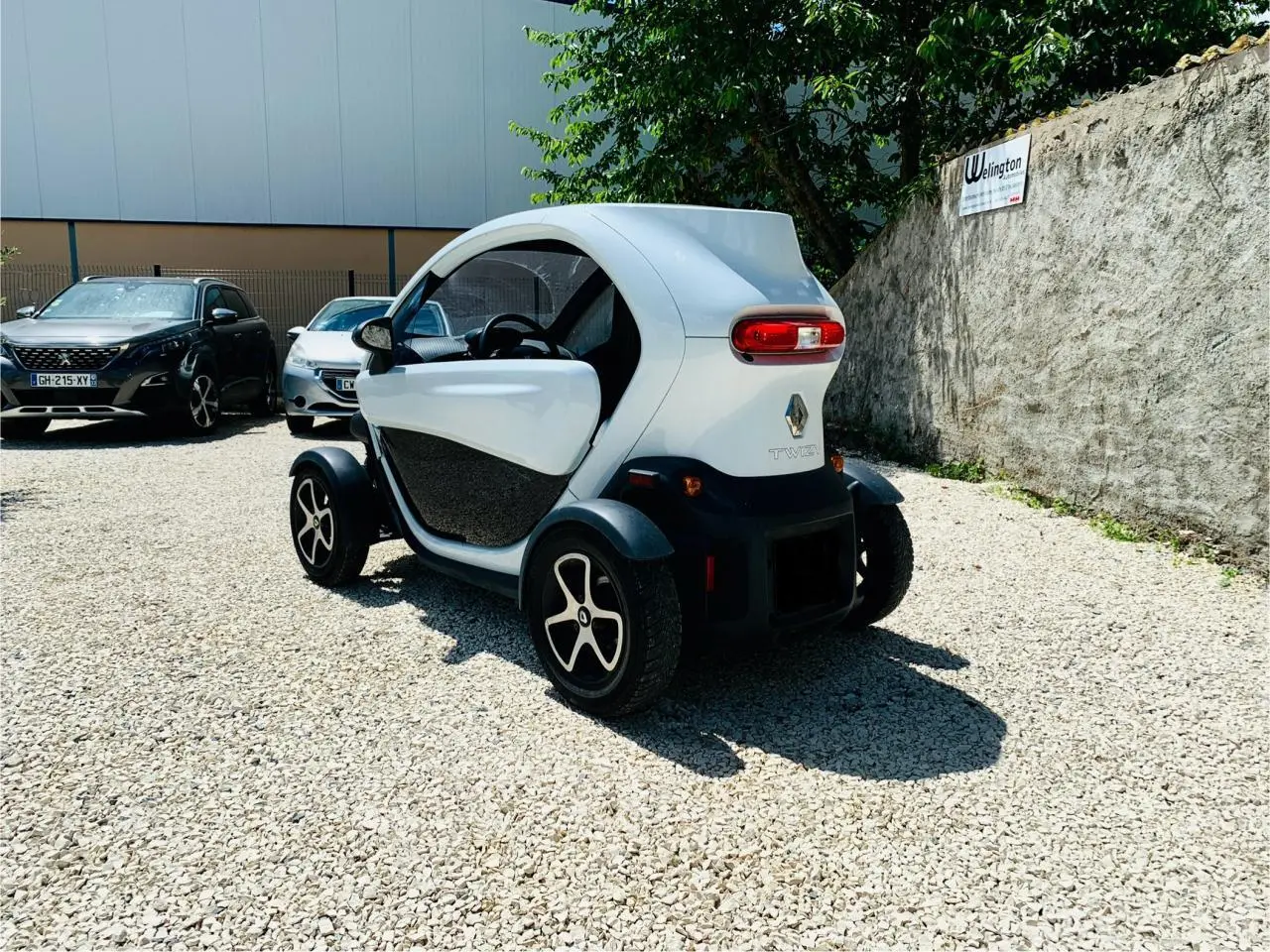 Vue 3/4 arrière droite d'une Renault Twizy blanche électrique garée sur un sol gravillonné, avec portes ouvertes.