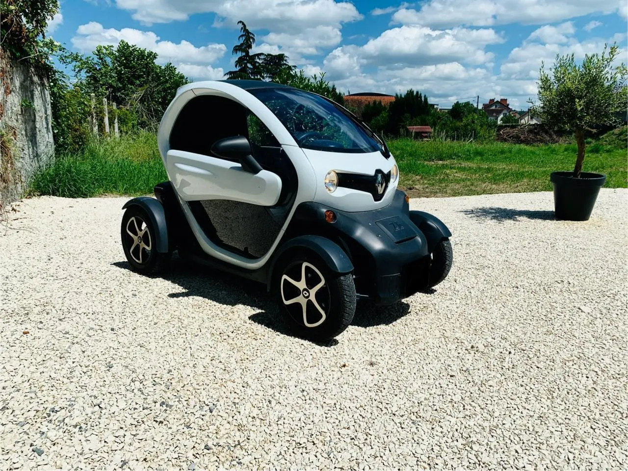 Vue 3/4 avant droite d'une Renault Twizy blanche électrique garée sur un sol gravillonné sous un ciel bleu.