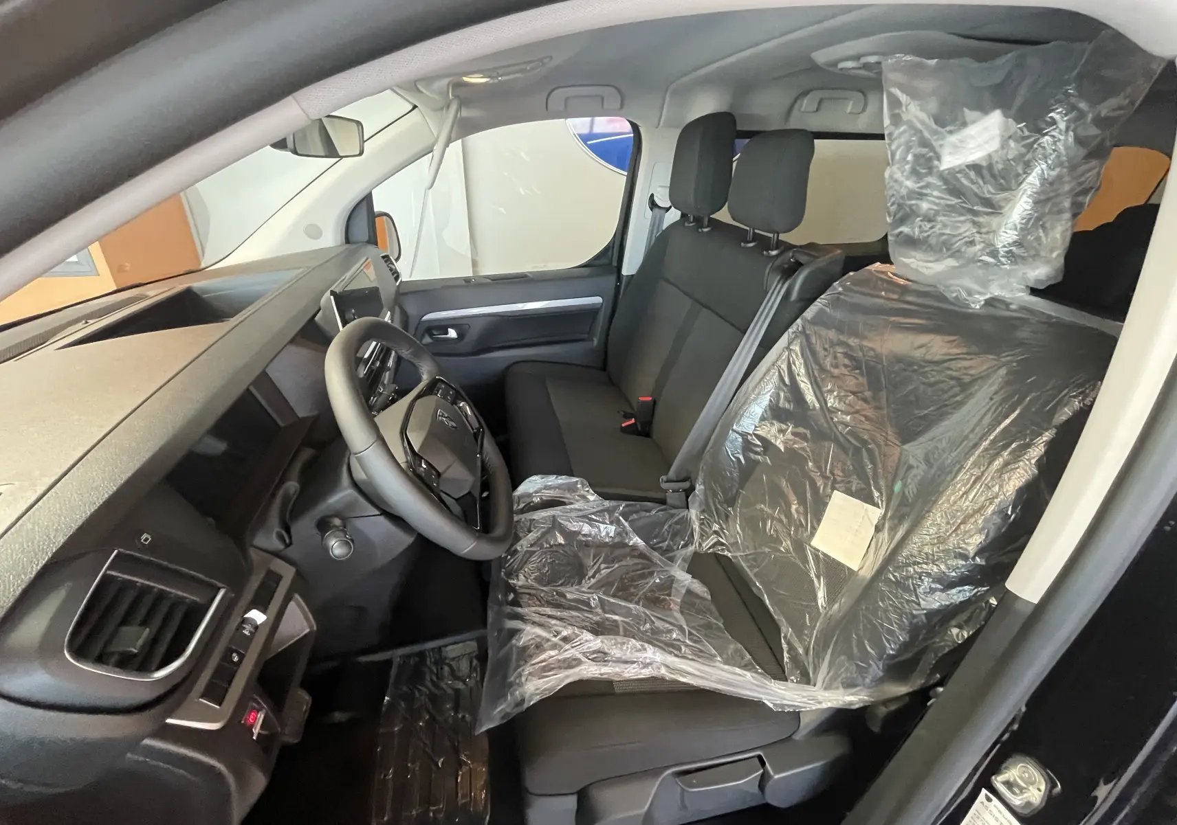 Vue intérieure côté conducteur du Peugeot Traveller noir 2026, montrant le volant cuir et les sièges avant protégés par un film plastique.