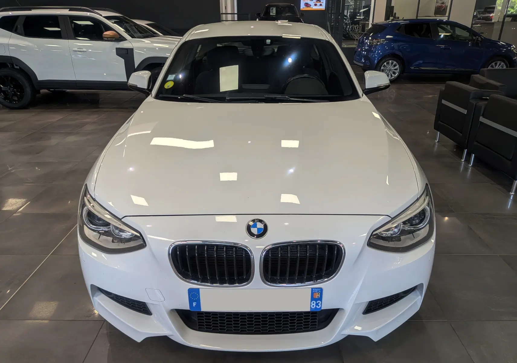 Vue frontale d'une BMW Série 1 F21 blanche Alpine M Sport 118d xDrive de 2014 en intérieur showroom.