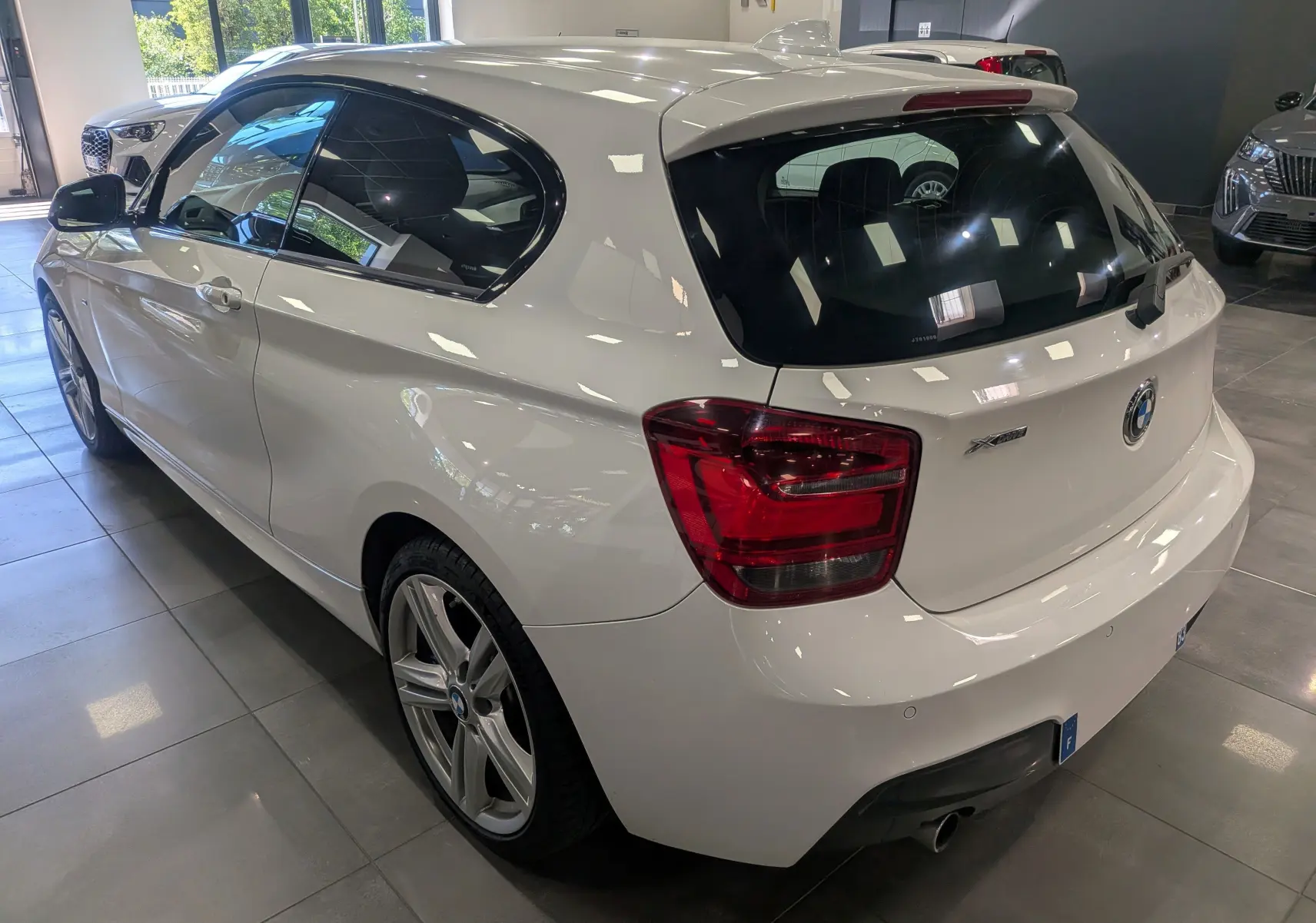 BMW Série 1 F21 blanc alpin vue 3/4 arrière droit, avec jantes alliage 18 pouces et feux arrière rouges.